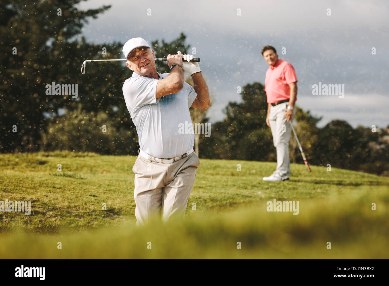 Golfeur Pro jouant sur le cours avec deuxième joueur debout à l'arrière à la recherche sur. L'homme de frapper la balle d'un bunker de sable au cours de la partie. Banque D'Images