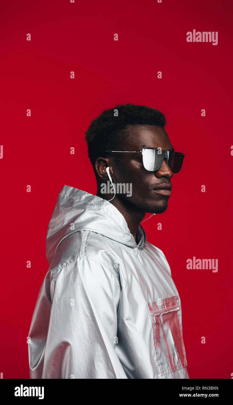 Homme africain en tenue futuriste contre fond rouge. L'Homme à lunettes élégantes, argent et capuche écouteurs. Banque D'Images