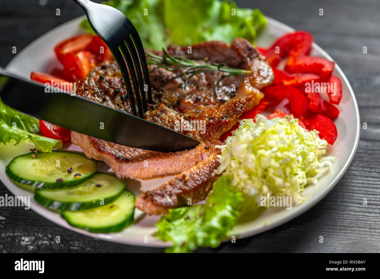 Couper avec le couteau et la fourchette de délicieuses grillades de viandes de porc et de sel poivrons parsemées avec légumes Banque D'Images