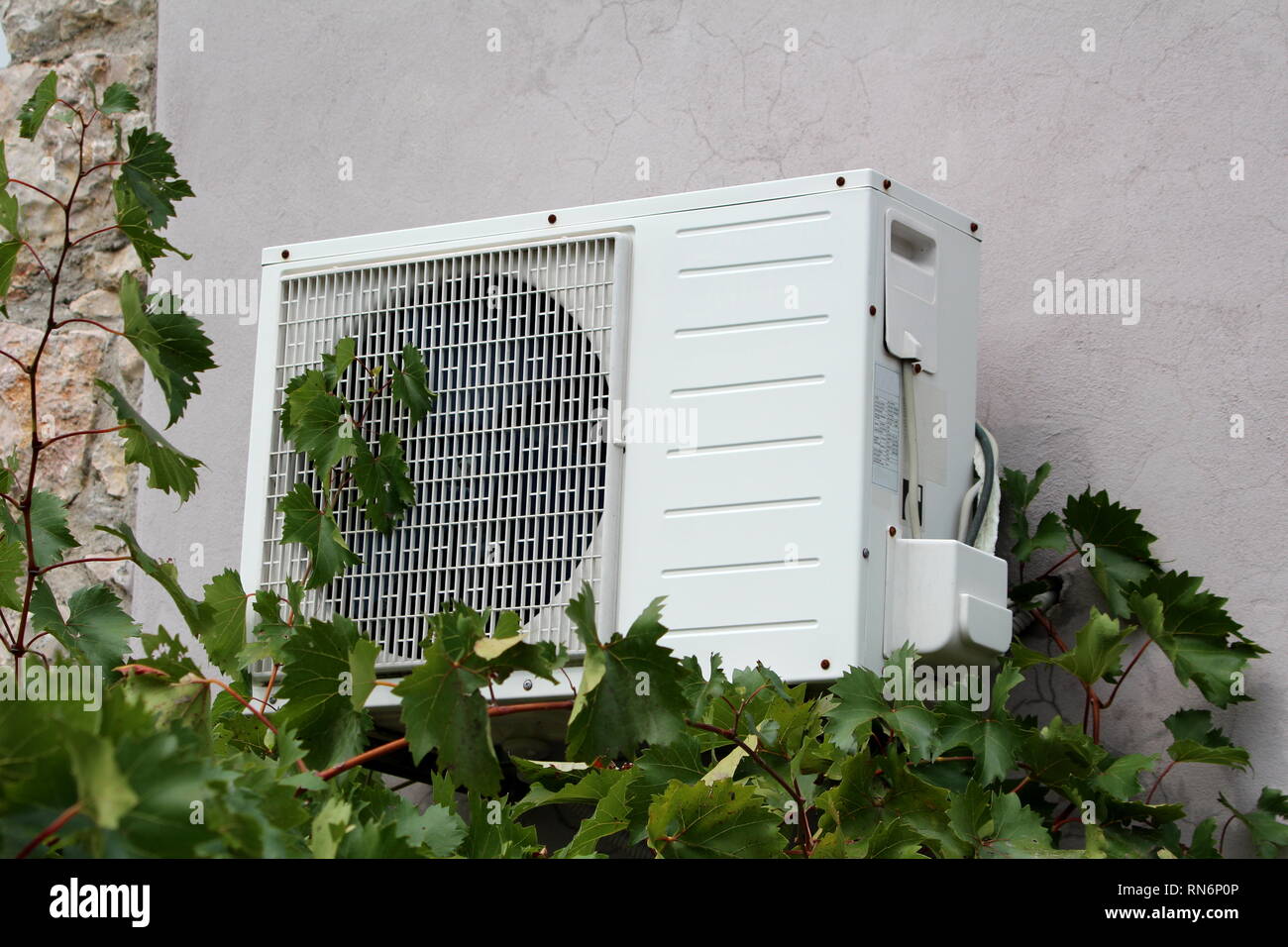 Fixée au mur à l'extérieur de l'unité AC climatiseur monté sur le mur arrière fissuré carreaux en pierre avec une partie de la maison de famille en face de plants de vigne jardin dense Banque D'Images
