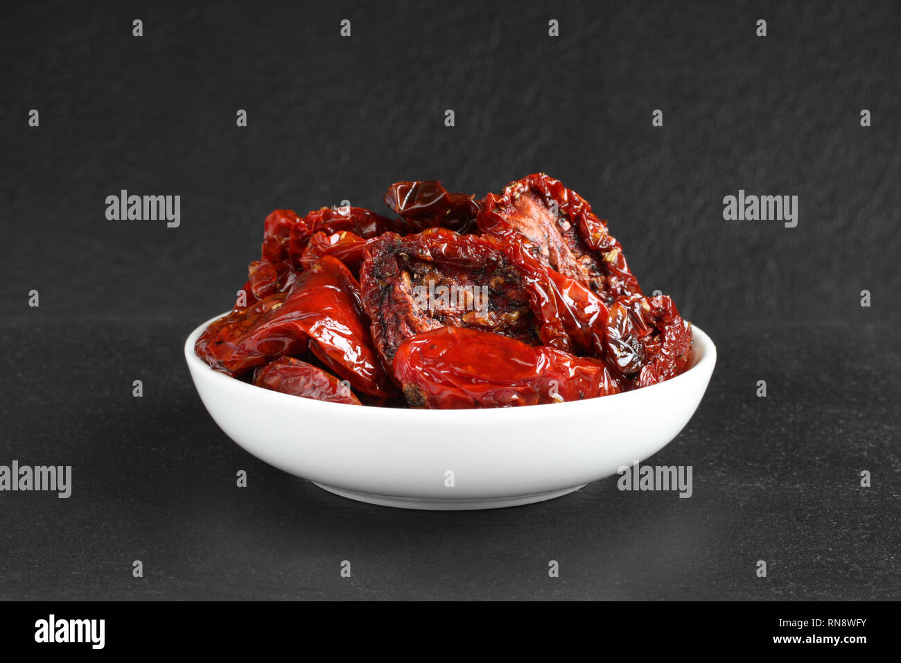 Tomates séchées dans l'huile de tournesol avec des épices (romarin, ail, basilic, poivre noir) dans un bol en céramique blanc, debout sur une pierre noire aga de surface Banque D'Images