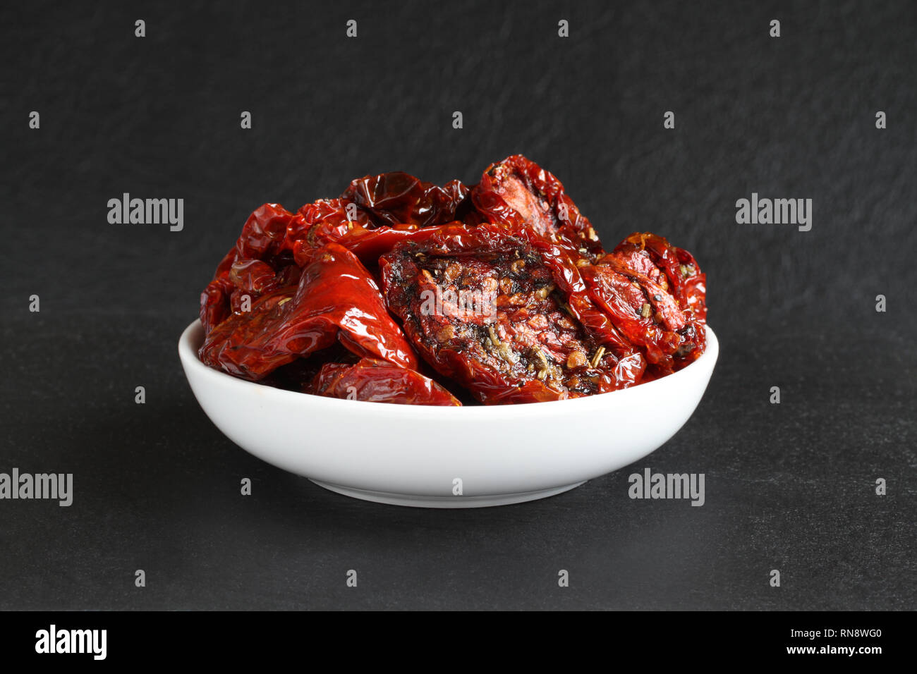 Tomates séchées dans l'huile de tournesol avec des épices (romarin, ail, basilic, poivre noir) dans un bol en céramique blanc, debout sur une pierre noire aga de surface Banque D'Images