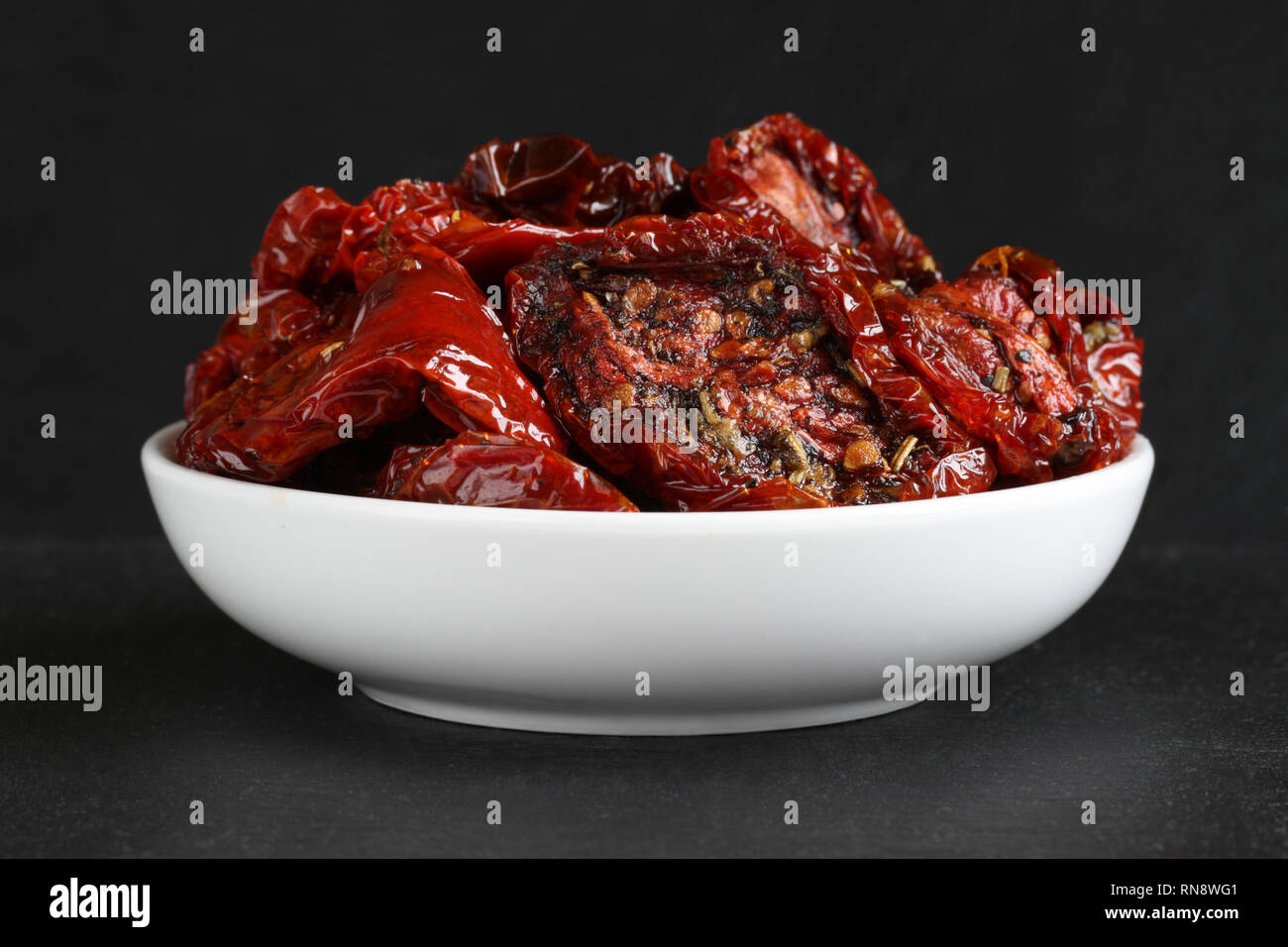 Tomates séchées dans l'huile de tournesol avec des épices (romarin, ail, basilic, poivre noir) dans un bol en céramique blanc, debout sur une pierre noire aga de surface Banque D'Images