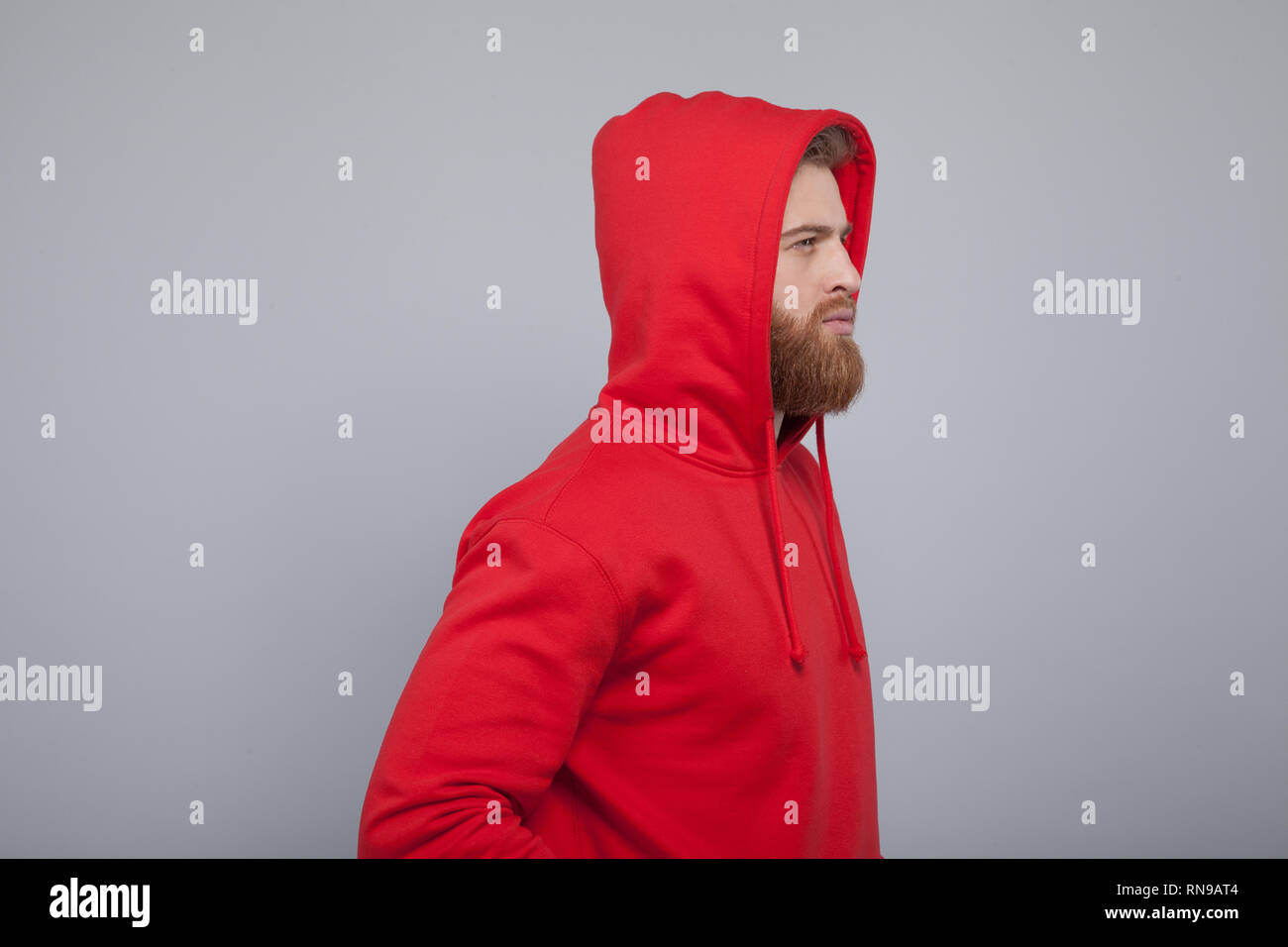 Beau jeune homme barbu tourné de profil portant des sweat rouge debout à l'arrière-plan gris regardant droit devant lui. Banque D'Images