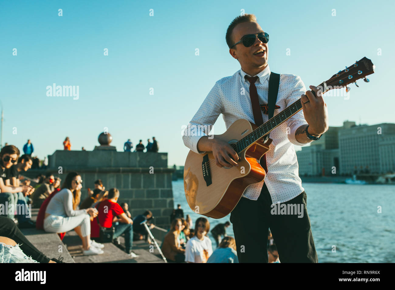 Moscou, Russie - 9 mai 2018 : jeune homme avec des lunettes de soleil, cravate et ruban de Saint-georges joue de la guitare à l'encontre de la rivière. Foule de gens assis et Banque D'Images