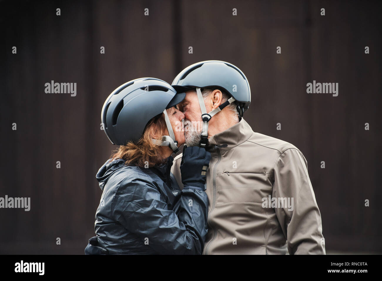 Senior couple avec casque de vélo debout à l'extérieur contre un arrière-plan sombre, s'embrasser. Banque D'Images