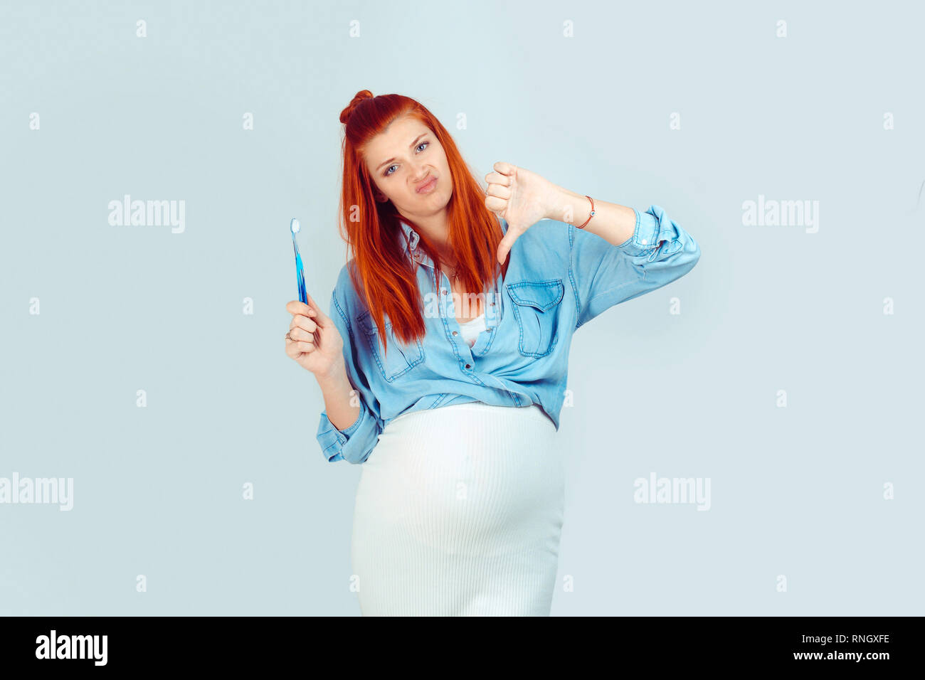 Jeune femme avec pregnant belly showing thumb par aversion de nouveau alors que la brosse à dents isolés sur fond bleu clair. Banque D'Images