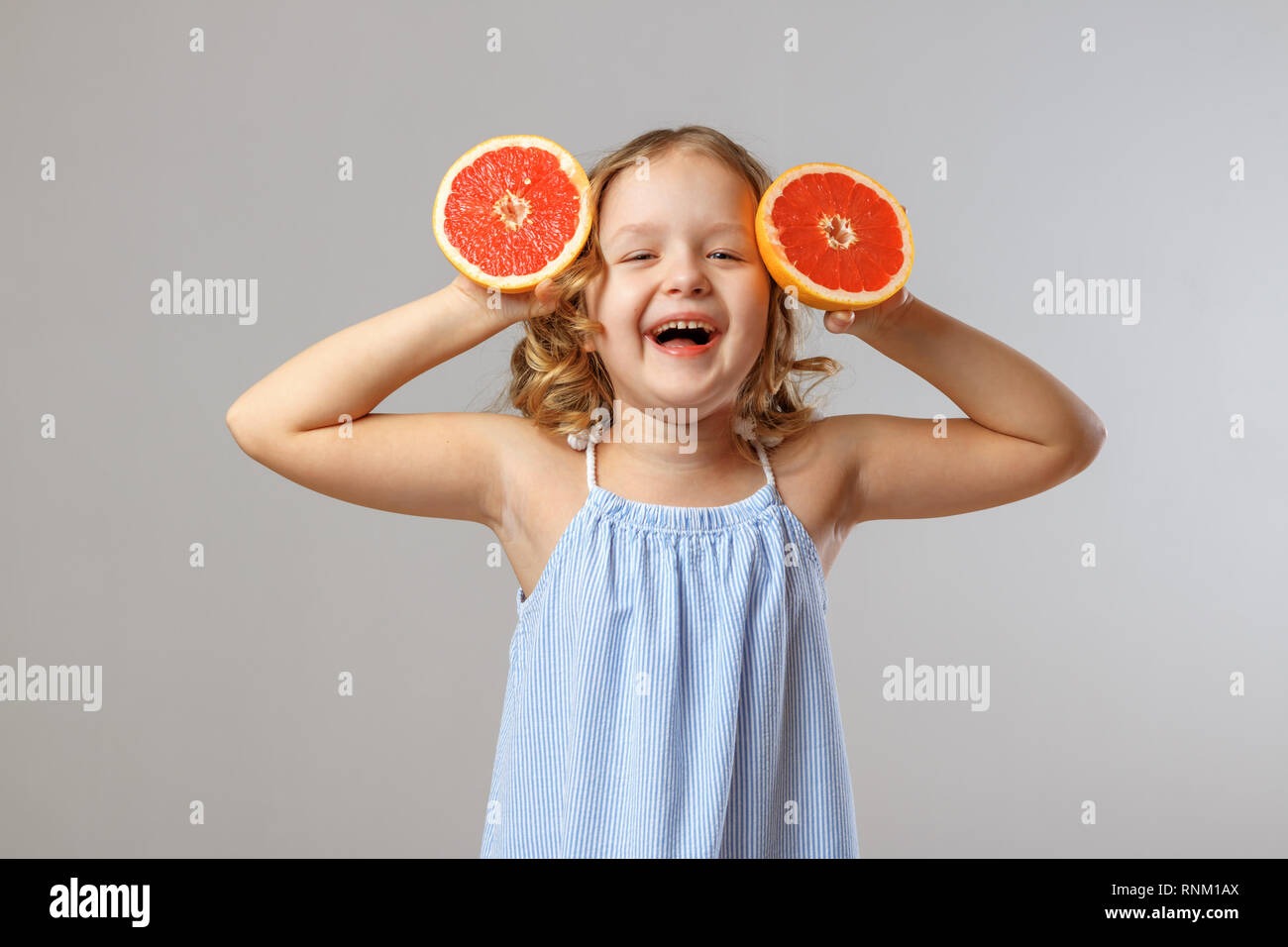 Petite fille joyeuse enfant rire et substituts moitiés de pamplemousse au lieu d'oreilles. Fond gris, studio Banque D'Images