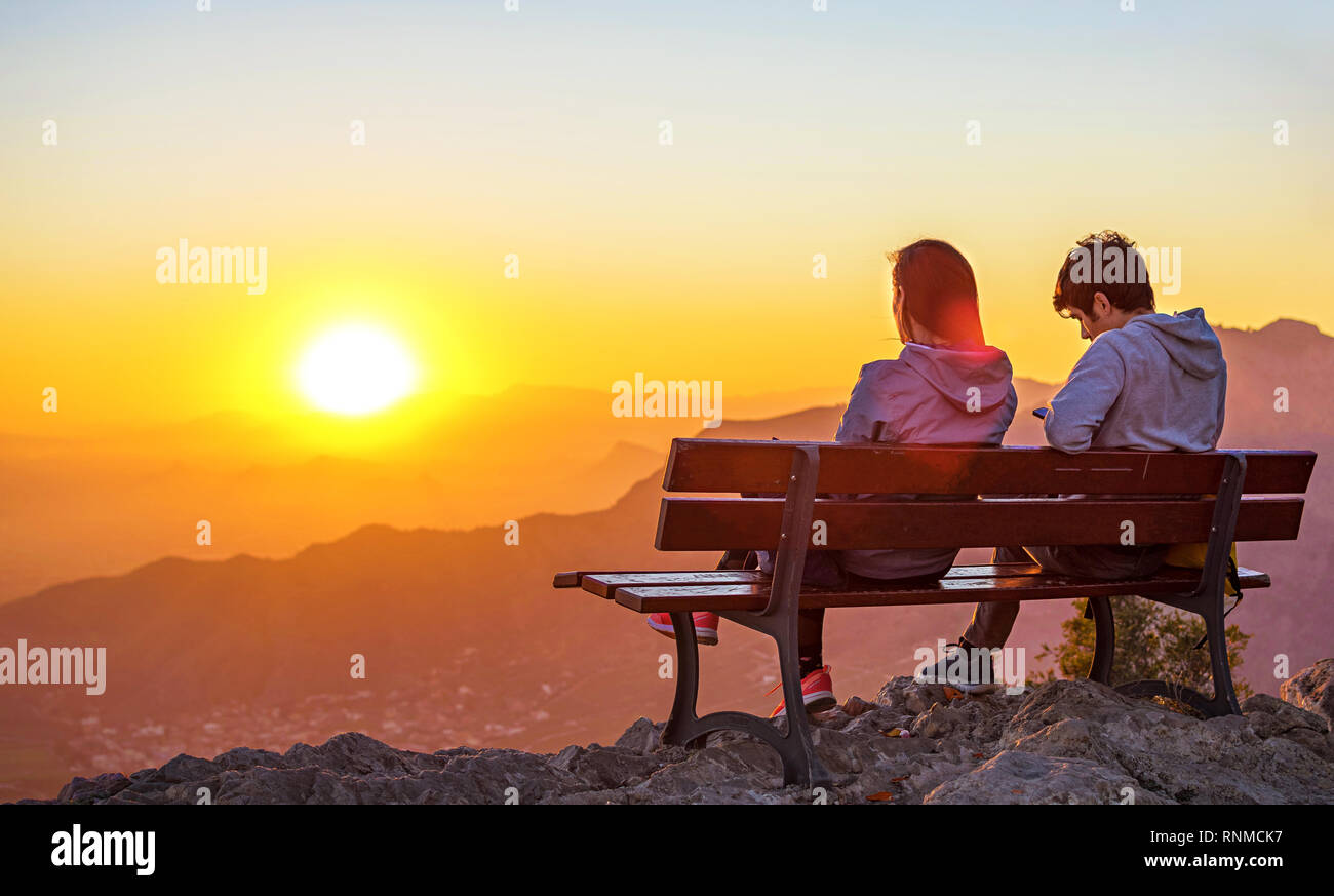 Deux personnes, en couple assis ensemble sur un banc au sommet d'une montagne à la recherche au coucher du soleil au cours de l'heure d'or. Banque D'Images