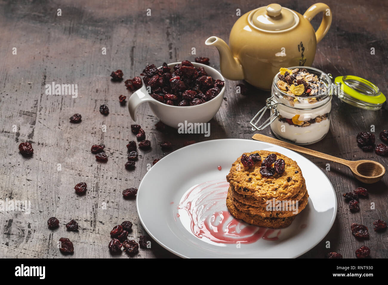 Photo horizontale avec plusieurs biscuits céréales mis sur plaque blanche. Confiture rouge est renversé sur la plaque. Canneberges rouges secs sont en blanc tasse et aro renversé Banque D'Images