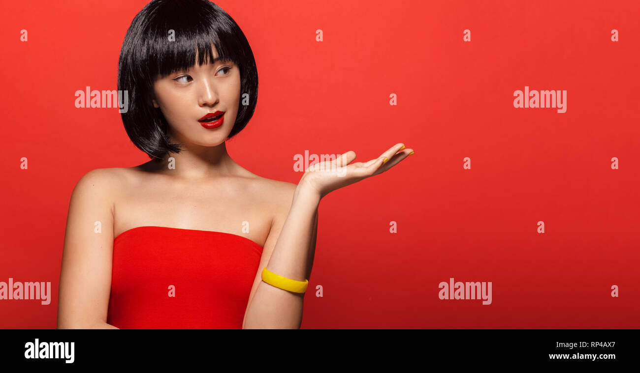Portrait of attractive young asian woman avec les cheveux courts en montrant l'espace de copie. Modèle féminin élégant vous montrant un produit sur fond rouge. Banque D'Images