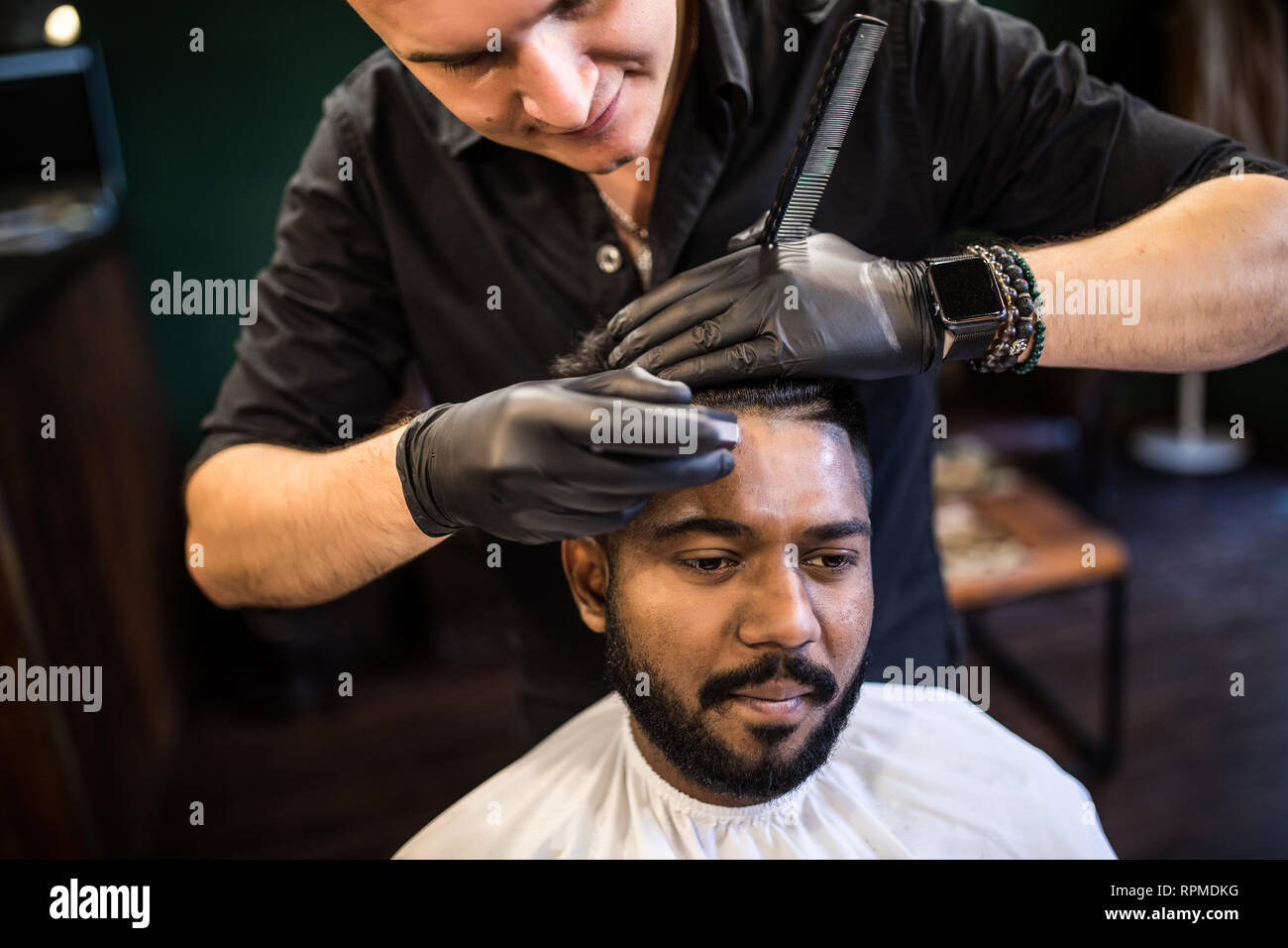 Jeune homme barbu se coupe par coiffure avec rasoir électrique tout en restant assis dans fauteuil de barbier Banque D'Images