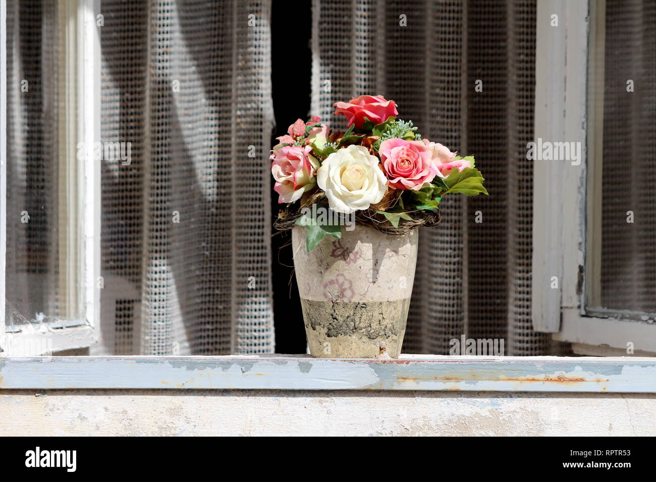 Vase en céramique cassée en partie avec des roses en plastique de différentes couleurs sur le dessus du joint de fenêtre avec les fenêtres ouvertes et le rideau en arrière-plan Banque D'Images