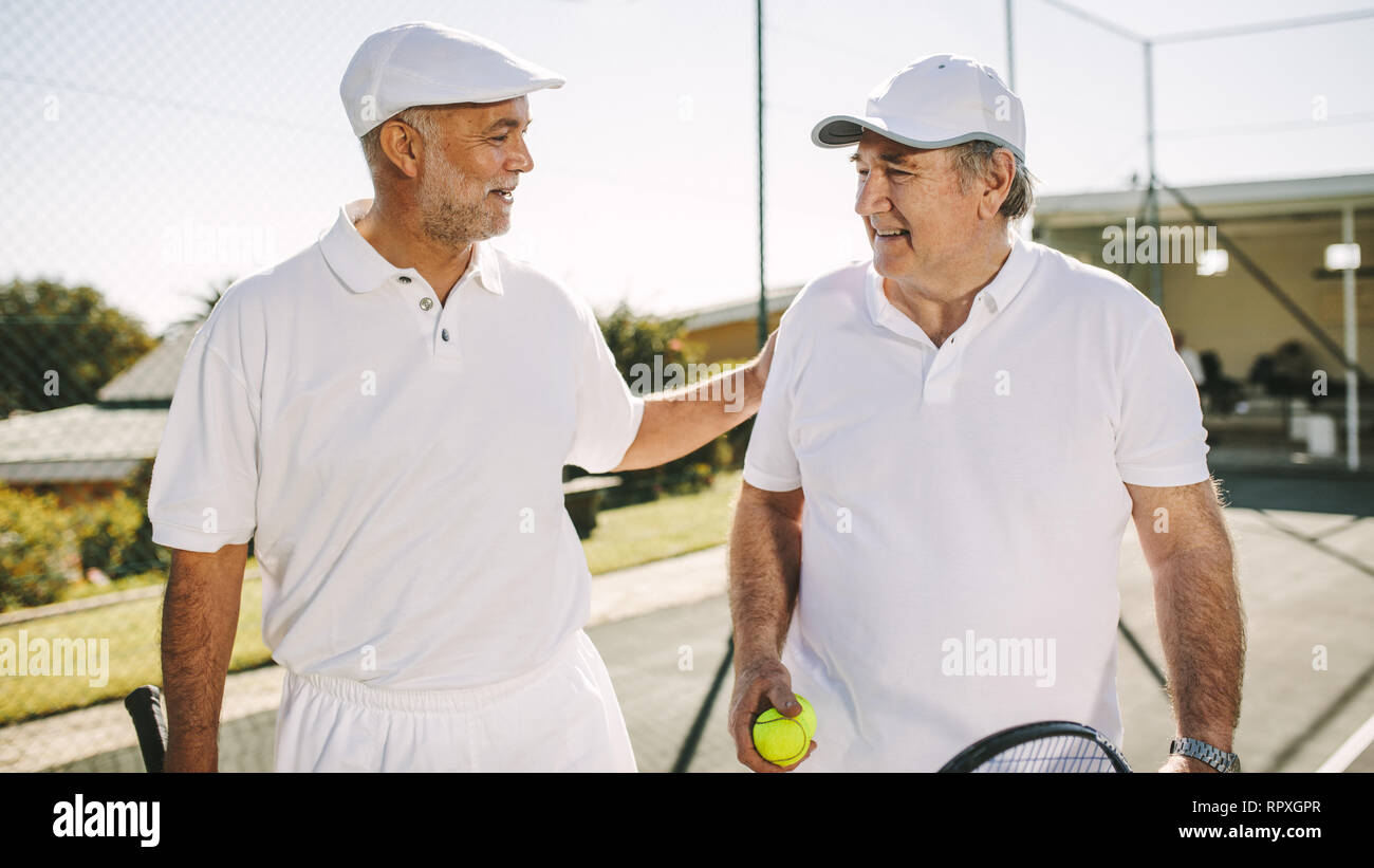 Les hommes de marcher sur un court de tennis pendant un match de tennis. Smiling senior hommes à porter des tennis de parler les uns aux autres la tenue des raquettes de tennis et de balle. Banque D'Images