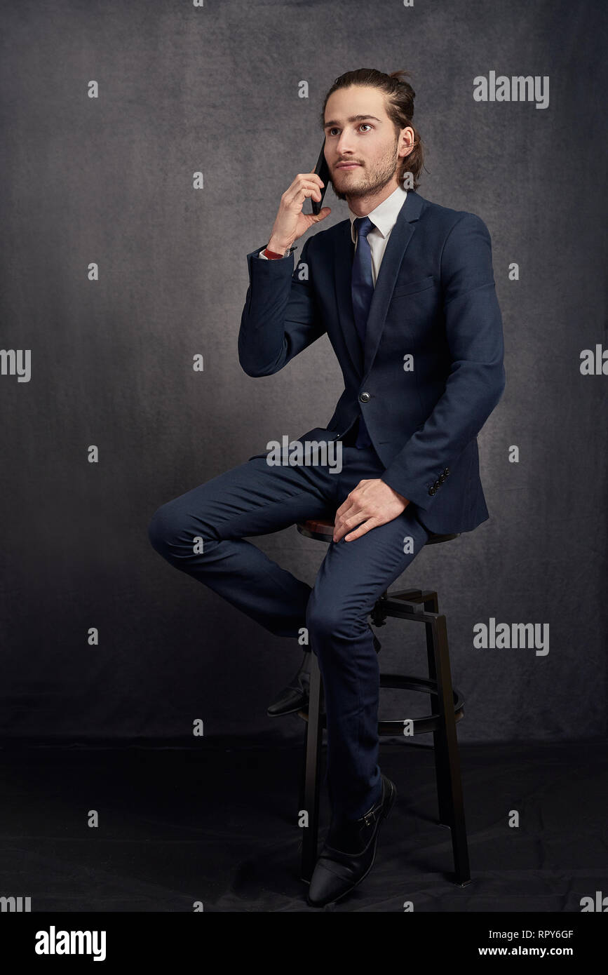 Jeune homme intelligent dans un élégant costume et cravate assis sur un tabouret à l'aide d'un téléphone mobile à l'écoute de l'appel avec une expression sérieuse sur dark gr Banque D'Images