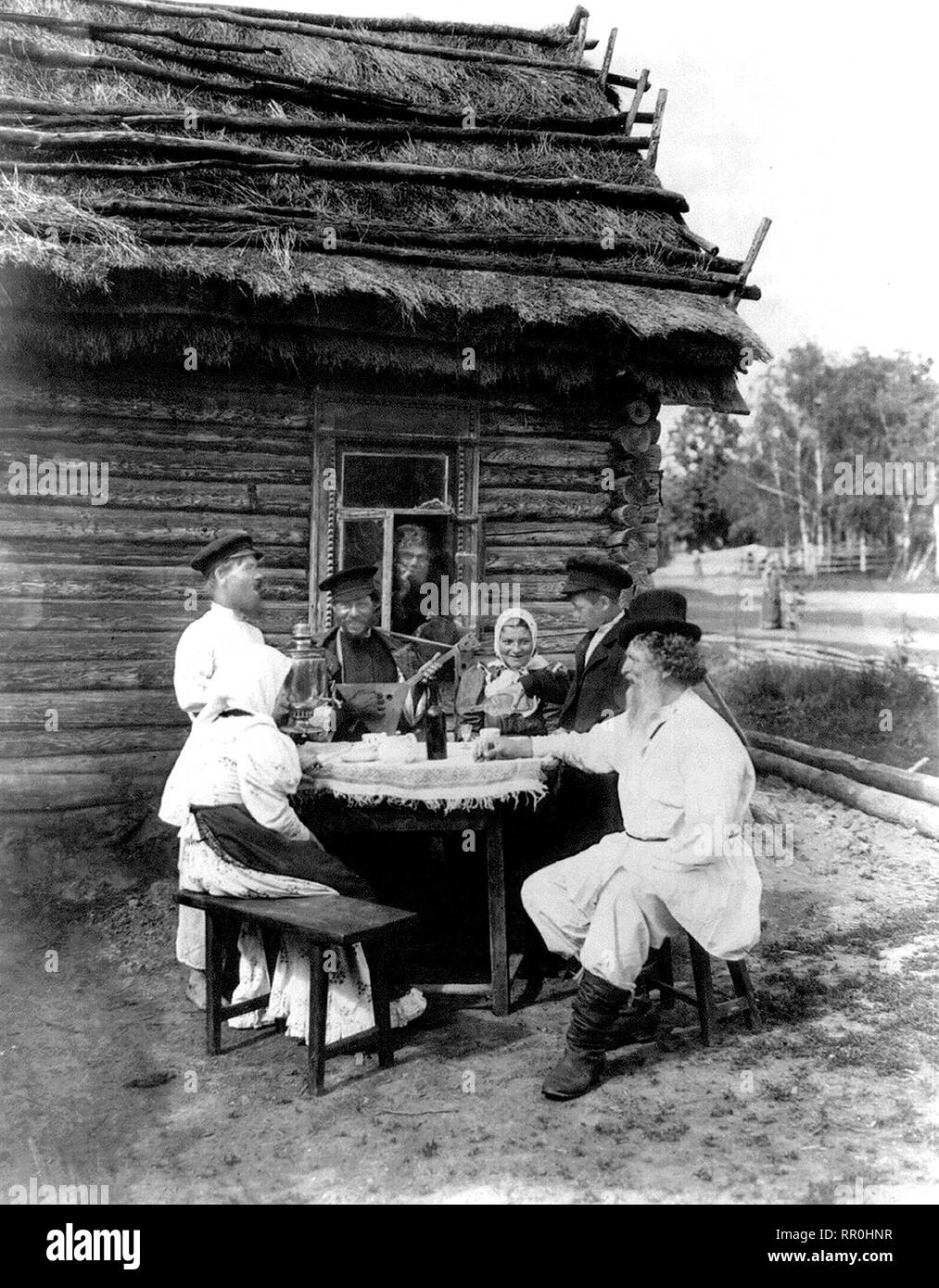 Genre scène vue inclut l'homme avec de la balalaïka, samovar sur la table, verser à boire, l'homme garçon par fenêtre et d'autres assis à table à l'extérieur d'une maison au toit de chaume. S paysan russe 1875 Banque D'Images