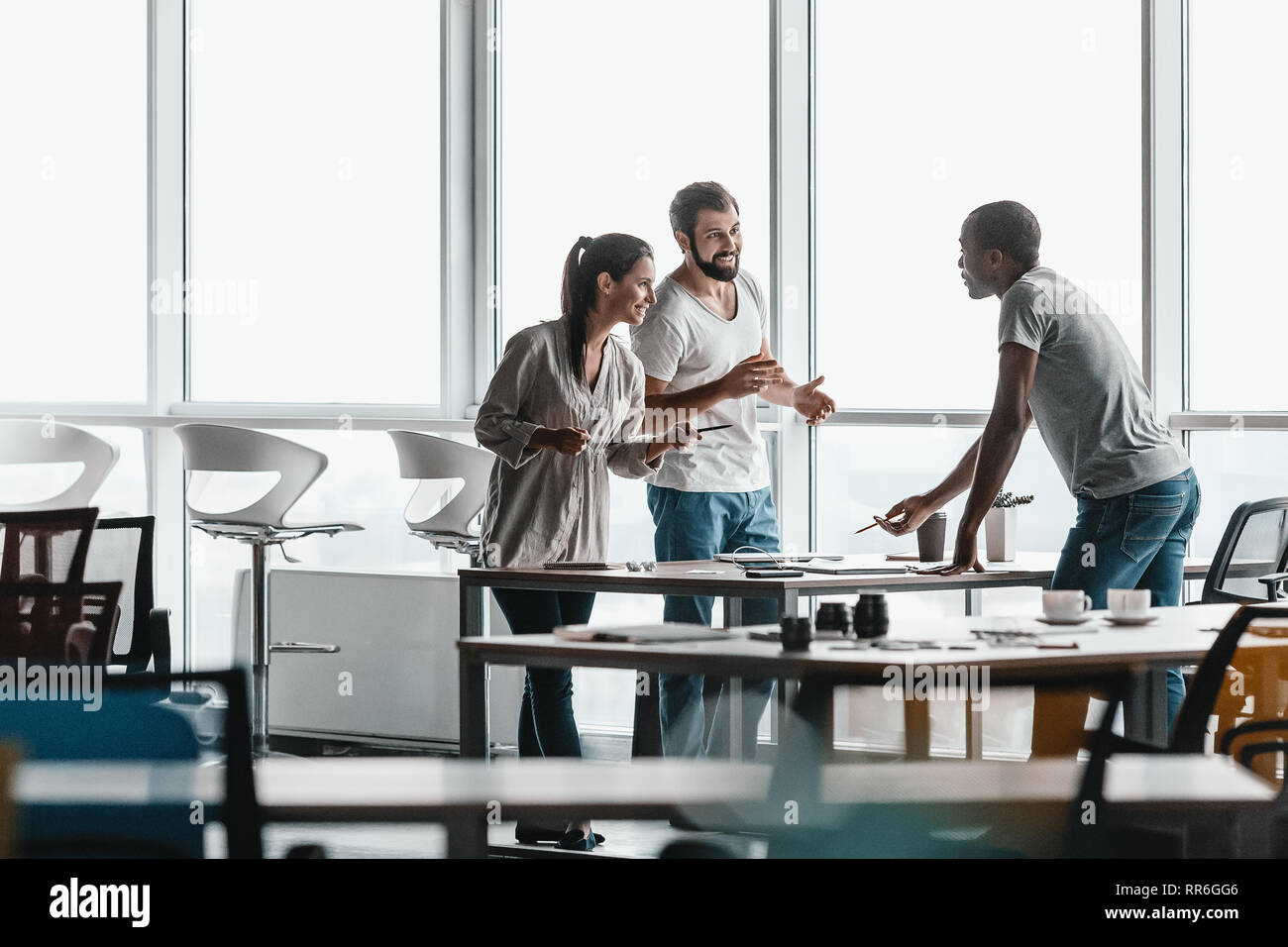 Trois hommes d'affaires dans les vêtements de la conversation tout en comité permanent par table en bois clair et moderne, à l'office. Ils sont souriants et regarder tout en Banque D'Images