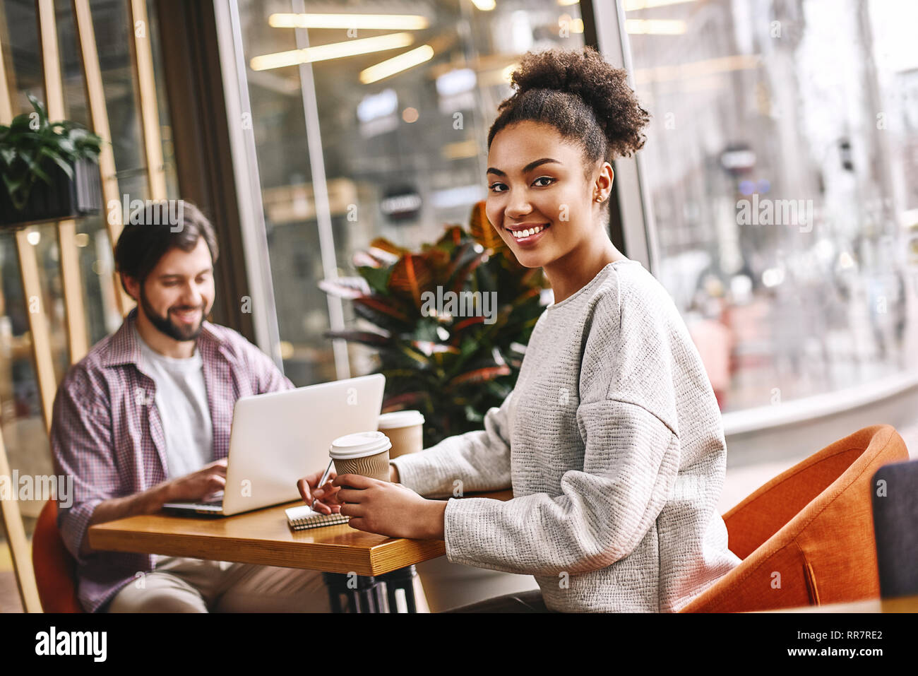 Joyeux deux collègues travaillent assis plus de 24. Man barbu en utilisant son ordinateur portable et attrayant african american woman smiling to camera, holding coffee et l'écriture de nouvelles idées. Style décontracté. Concept de la réussite Banque D'Images