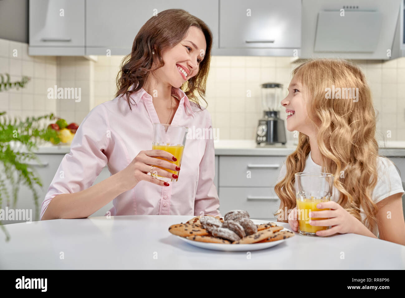 Jeune fille joyeuse et woman sitting at table dans la cuisine. Belle mère et belle fille de boire du jus d'orange et de manger des biscuits. Une jeune femme Banque D'Images