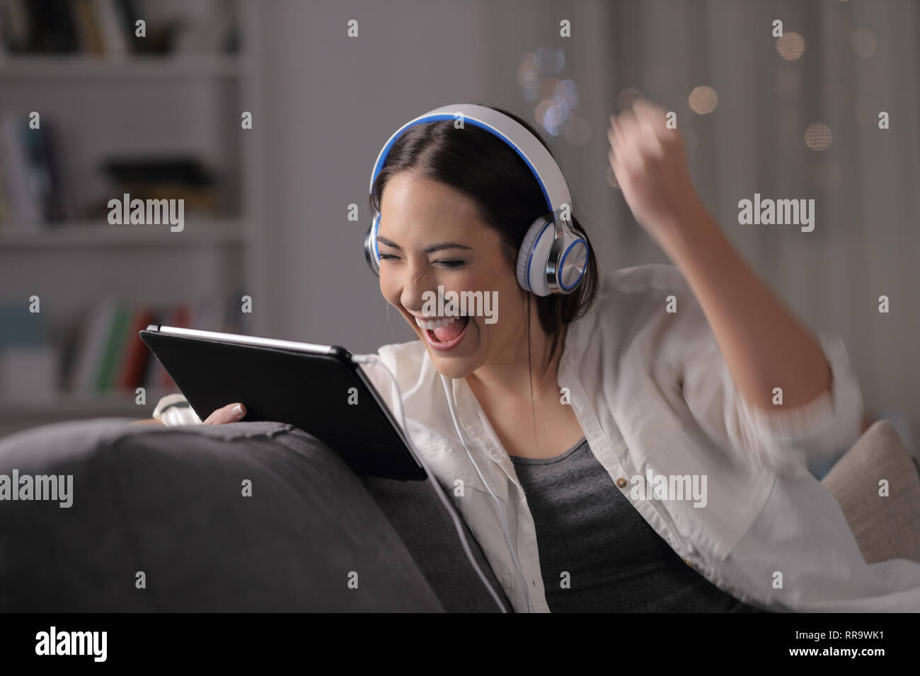 Excited woman écoute et regarde media sur tablet assis sur un canapé dans la nuit à la maison Banque D'Images
