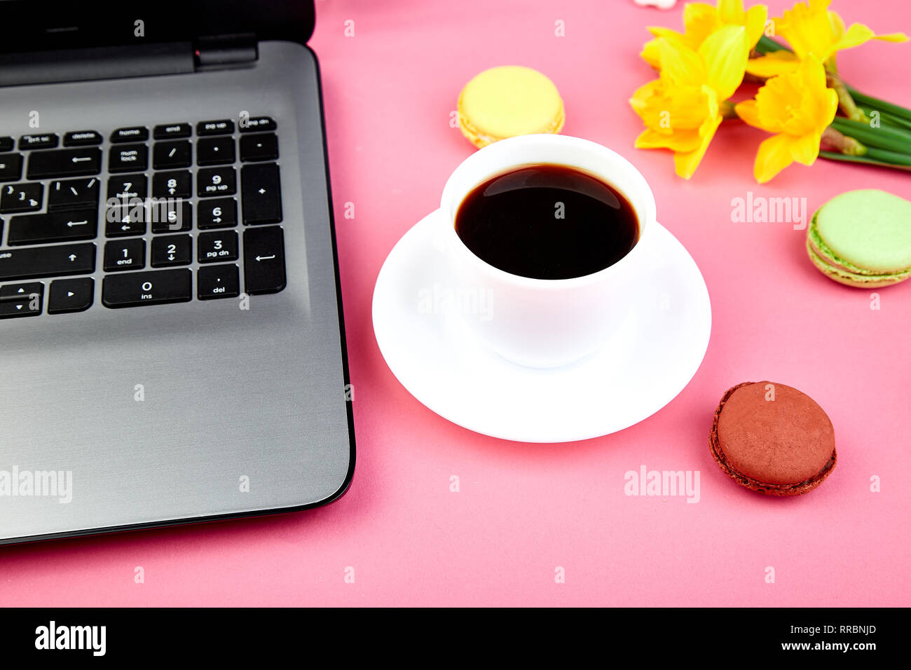 Femme ou espace de travail féminin avec ordinateur portable, portable, tasse de café, macarons, et fleurs narcisse sur table rose Banque D'Images