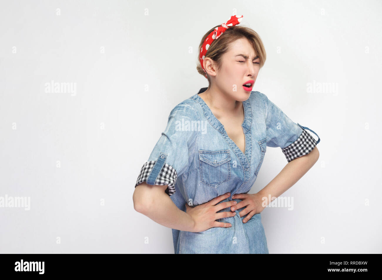 La douleur d'estomac ou de période. Portrait de la belle jeune femme en chemise en denim bleu décontracté avec un miroir, bandeau rouge debout et tenant son ventre douloureux. Banque D'Images