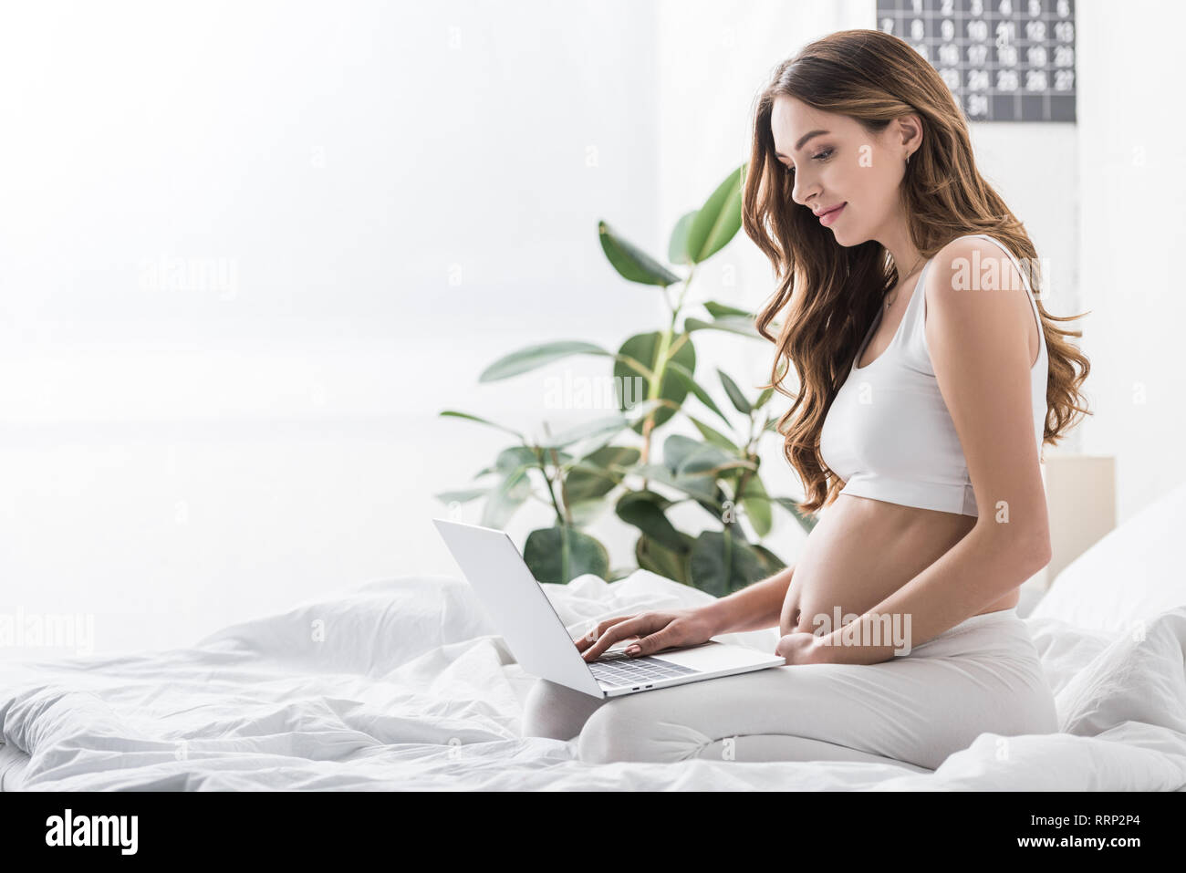 Charmante femme enceinte en utilisant laptop while sitting on bed Banque D'Images
