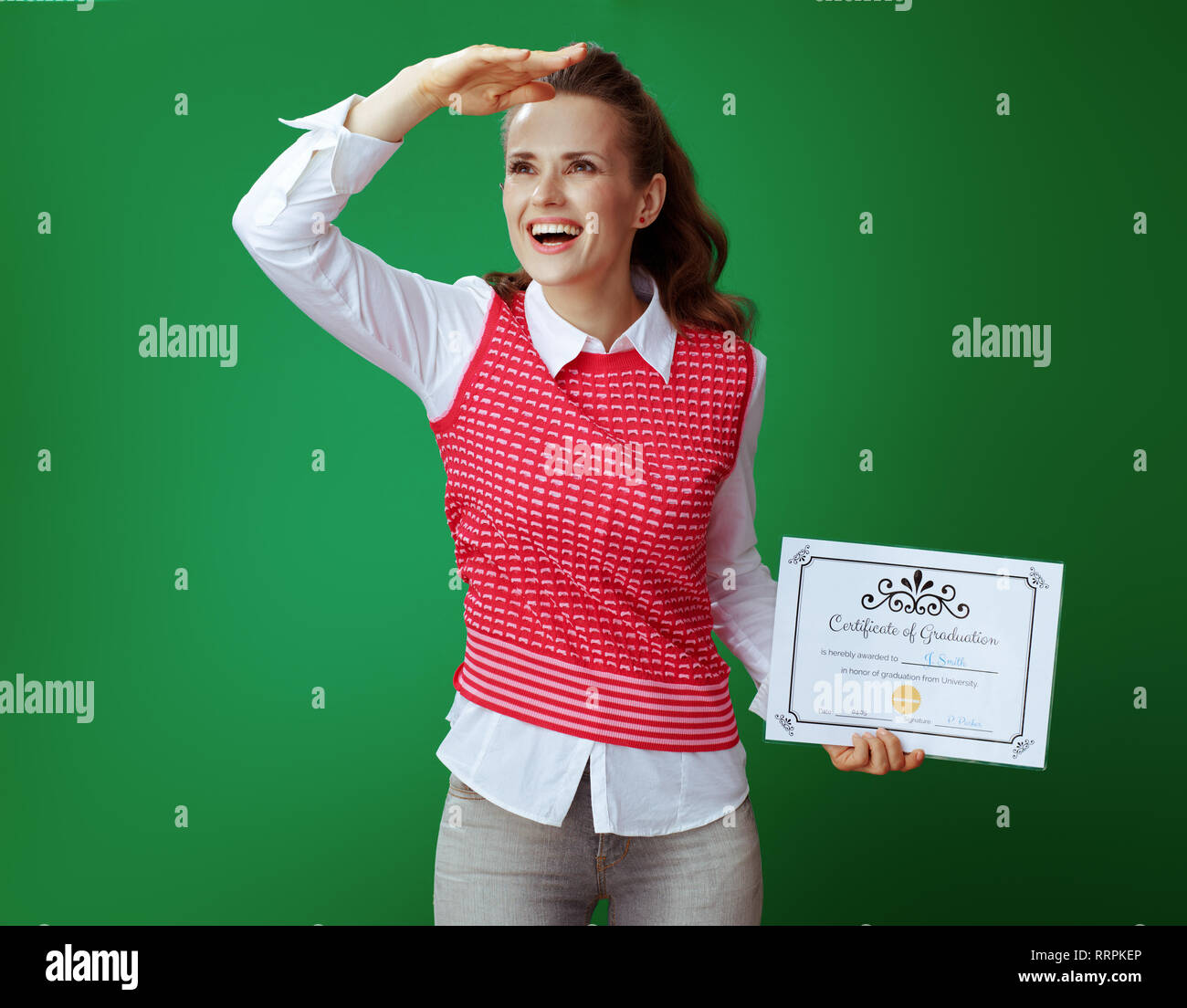 Heureux sain femme étudiant en gris jeans et t-shirt rose avec certificat de l'obtention du diplôme à la recherche dans la distance contre green chalkboard Banque D'Images