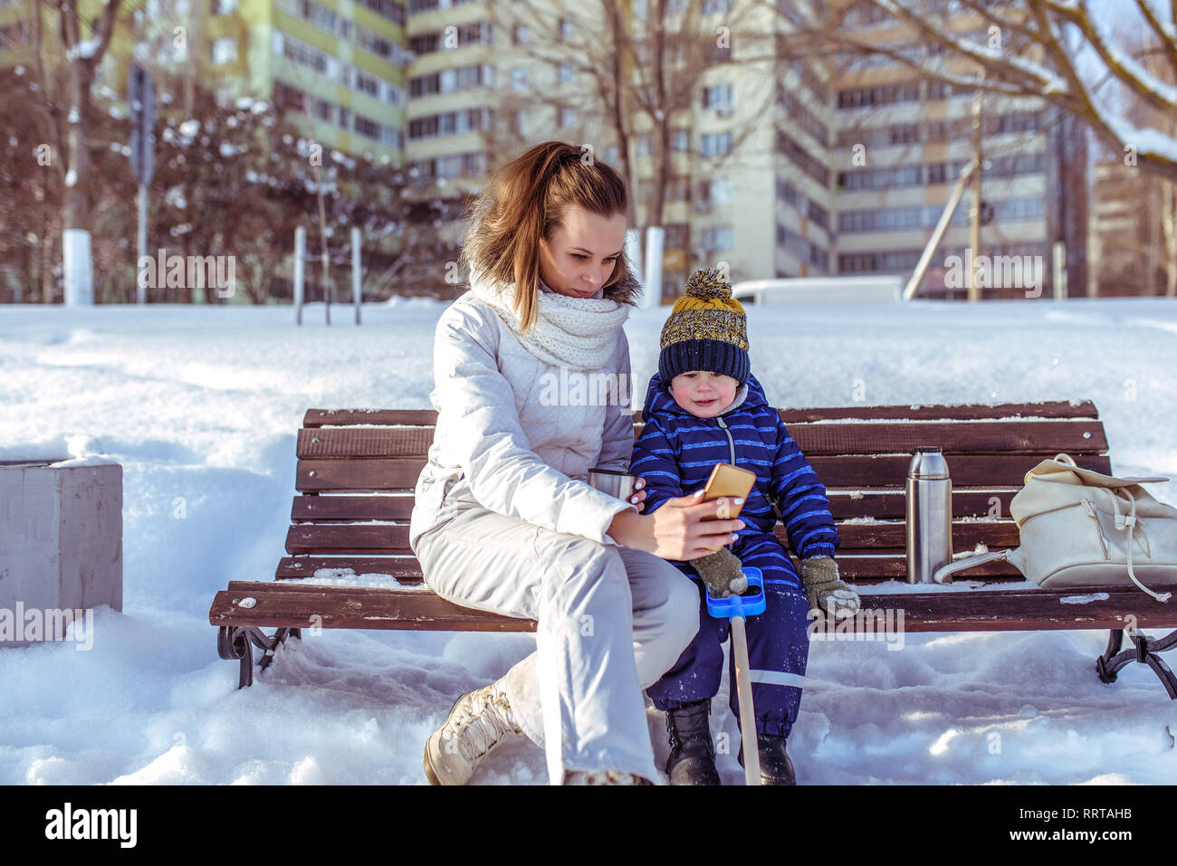 Petit garçon fils de 2-3 ans, assis banc en hiver ville. Femme maman de mains de téléphone portable, prendre des photos, de l'app en ligne dans les réseaux sociaux. Hiver Nature Banque D'Images