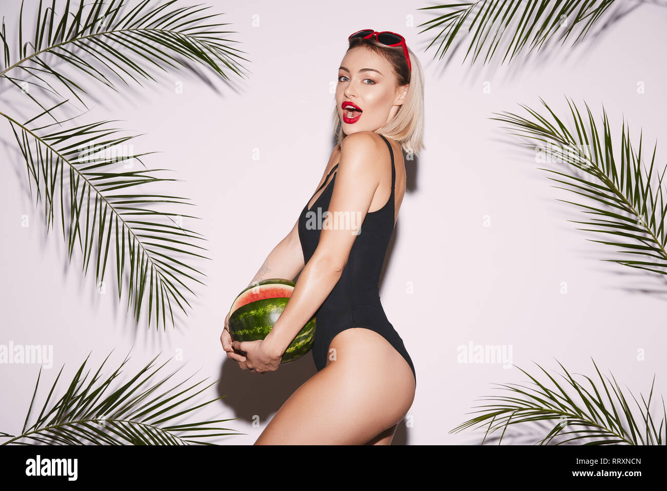 Belle fille avec des lèvres rouge vêtu de noir et des lunettes maillots de posing at white background studio avec palmiers, holding watermelon. Banque D'Images