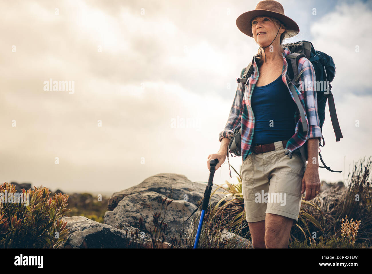 Senior woman wearing hat et son sac à dos de trekking dans la campagne tenant un pôle randonnée. Femme aventureux sur un sentier de randonnée sur une journée nuageuse. Banque D'Images