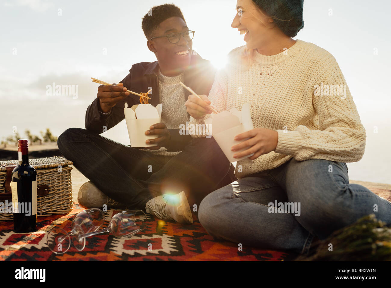 Couple Interracial on picnic eating noodles et souriant sur une journée ensoleillée. Smiling young man and woman sitting on picnic blanket ayant l'alimentation et à la Banque D'Images
