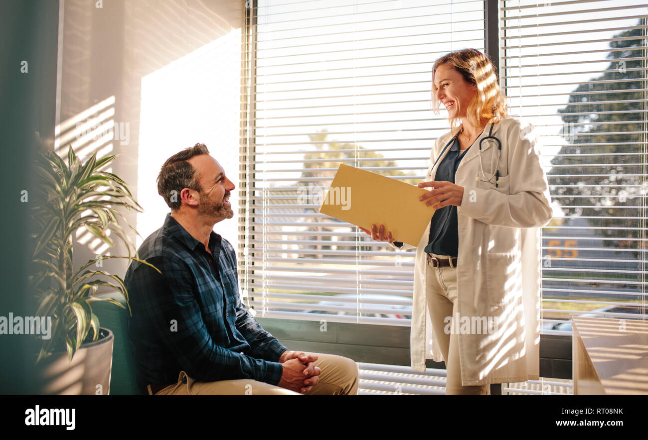 Smiling female doctor holding un fichier de rapport médical et parlant avec male patient assis sur un canapé à la réception de l'hôpital. Partage de bons services médicaux médecin t Banque D'Images