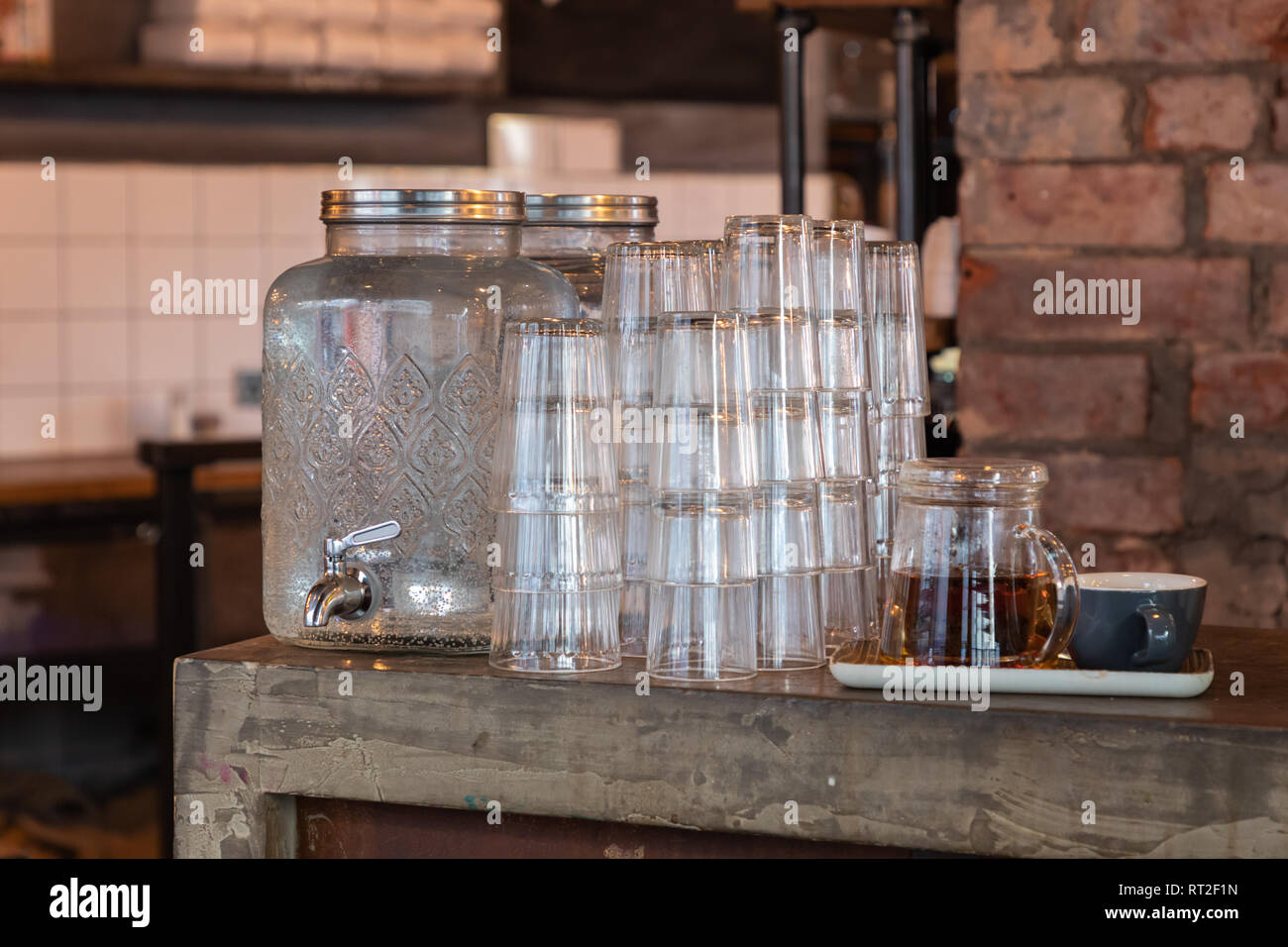 Un récipient d'eau en verre à motifs avec un robinet d'argent se dresse sur une table à côté de la pile de lunettes. À côté de lui est une tasse et une théière en verre. Banque D'Images