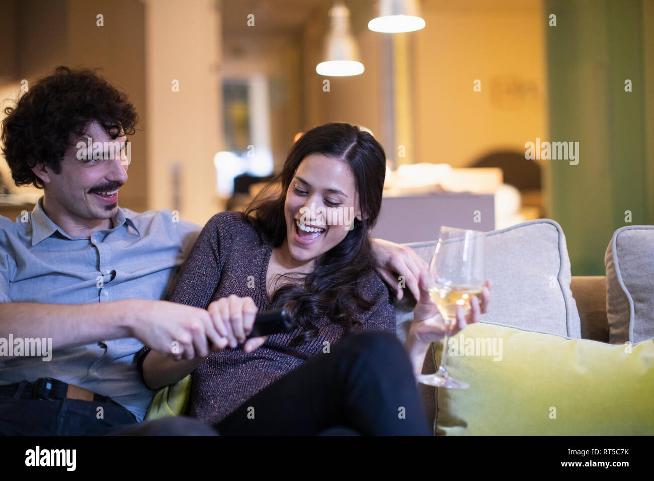 Playful couple lutte pour le contrôle à distance, de regarder la télévision et boire le vin blanc sur le canapé Banque D'Images