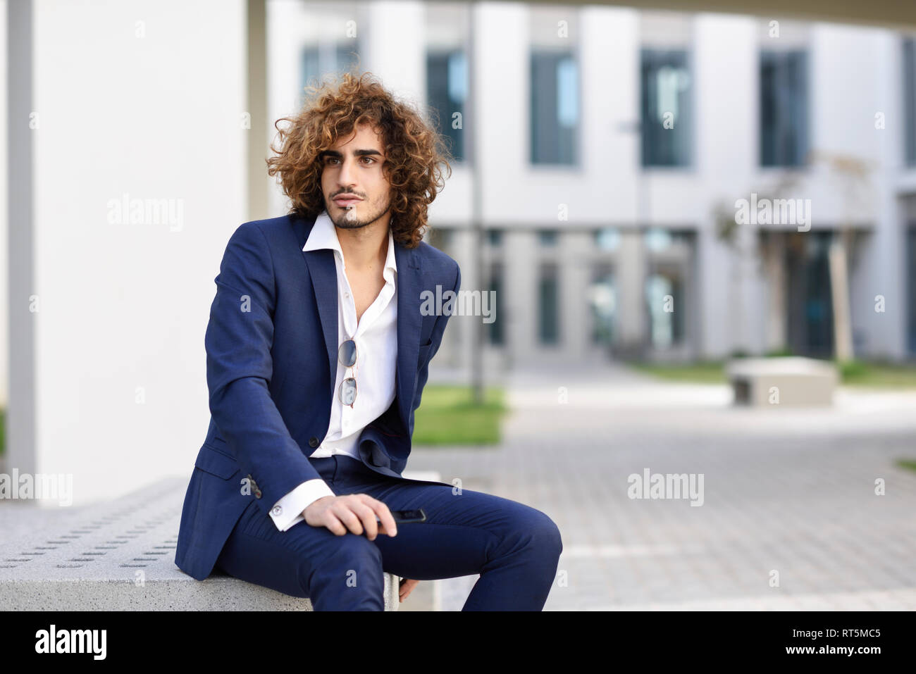 Portrait de jeune homme avec les cheveux bouclés portant costume bleu assis sur un banc à l'extérieur Banque D'Images