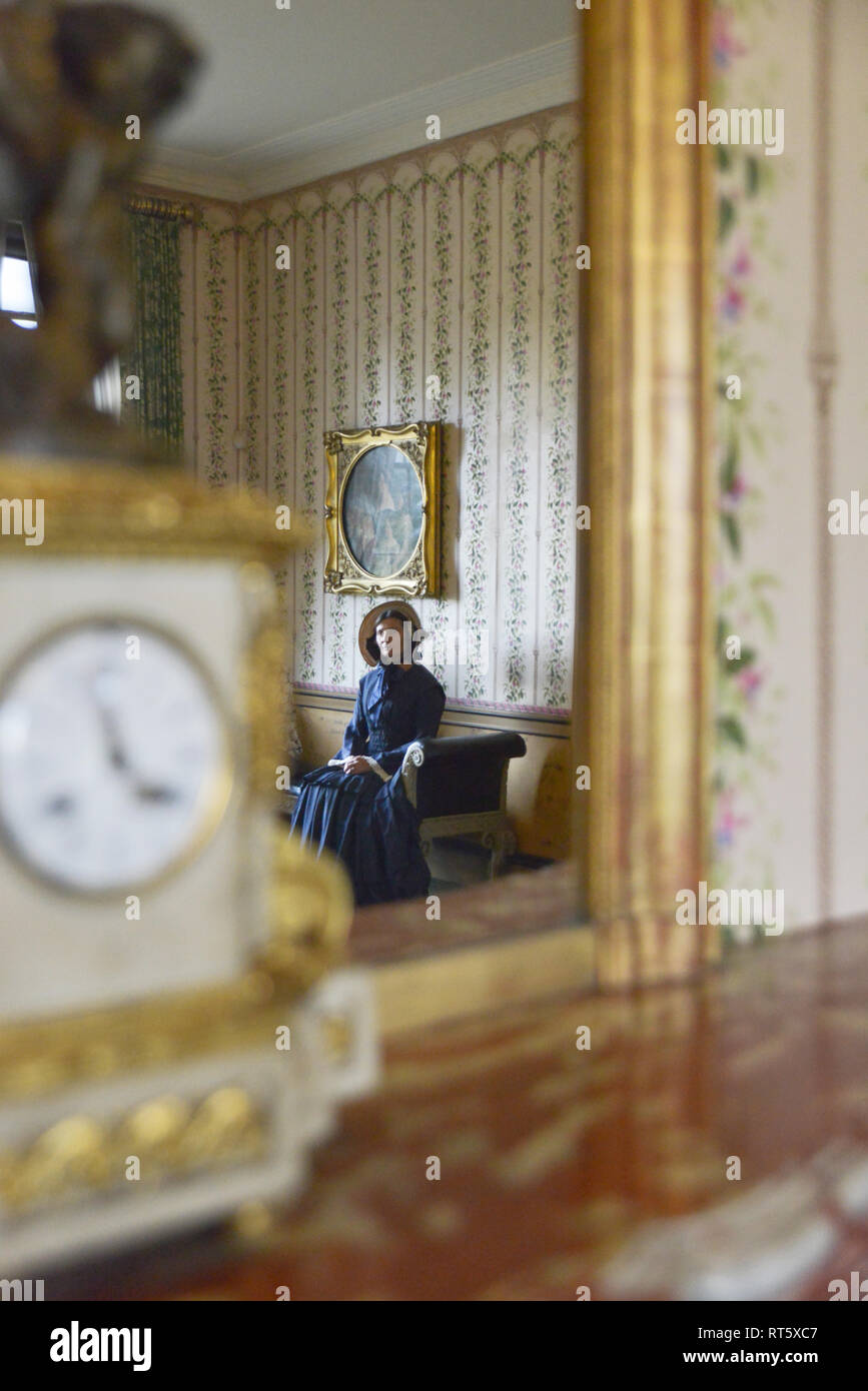 Une vieille femme se distingue par un style Victorien très cheminée décorée et regarde dans un miroir- il y a une horloge bronze doré d'or disant que c'est 4h00 à côté d'elle. Banque D'Images