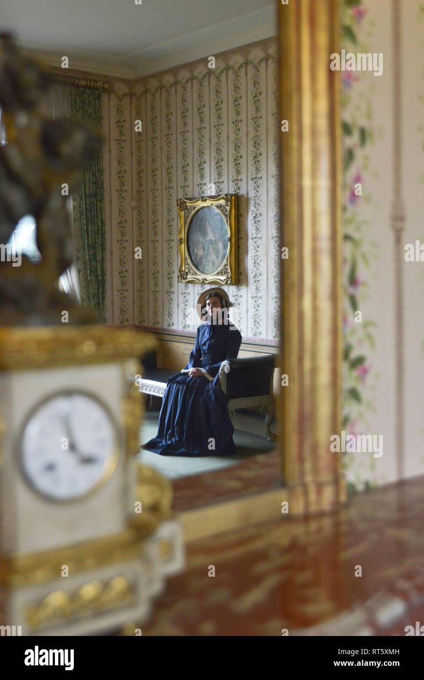 Une vieille femme se distingue par un style Victorien très cheminée décorée et regarde dans un miroir- il y a une horloge bronze doré d'or disant que c'est 4h00 à côté d'elle. Banque D'Images