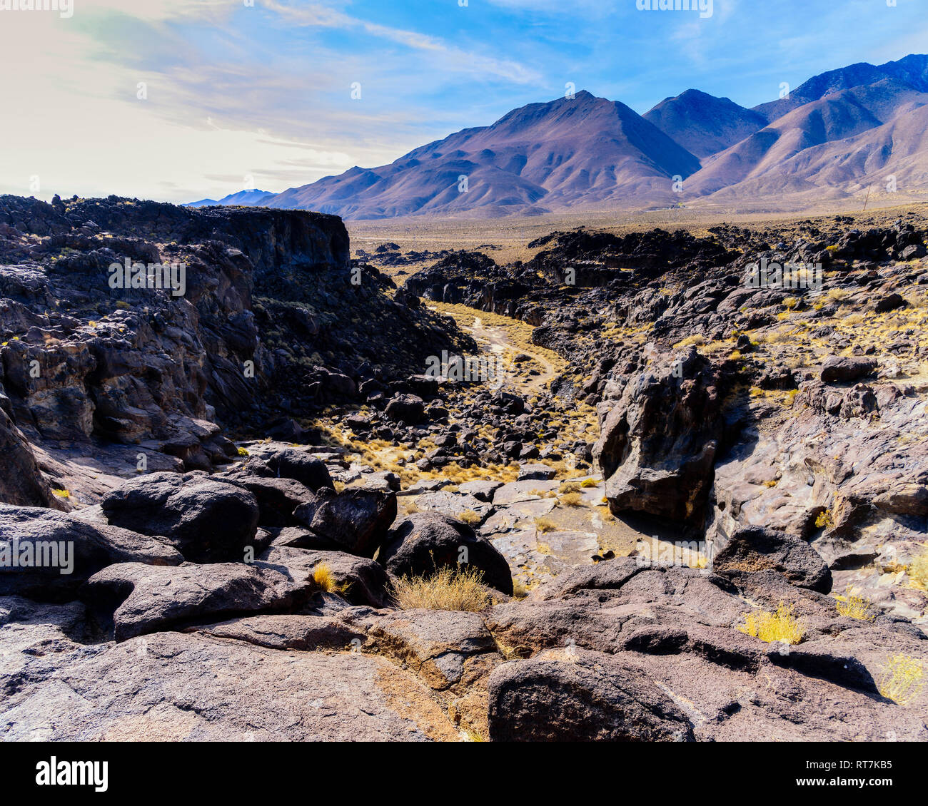 La recherche à travers un terrain accidenté à rocky canyon aux parois déchiquetées menant au désert aride vallée et les montagnes au-delà sous ciel bleu avec des nuages blancs. Banque D'Images