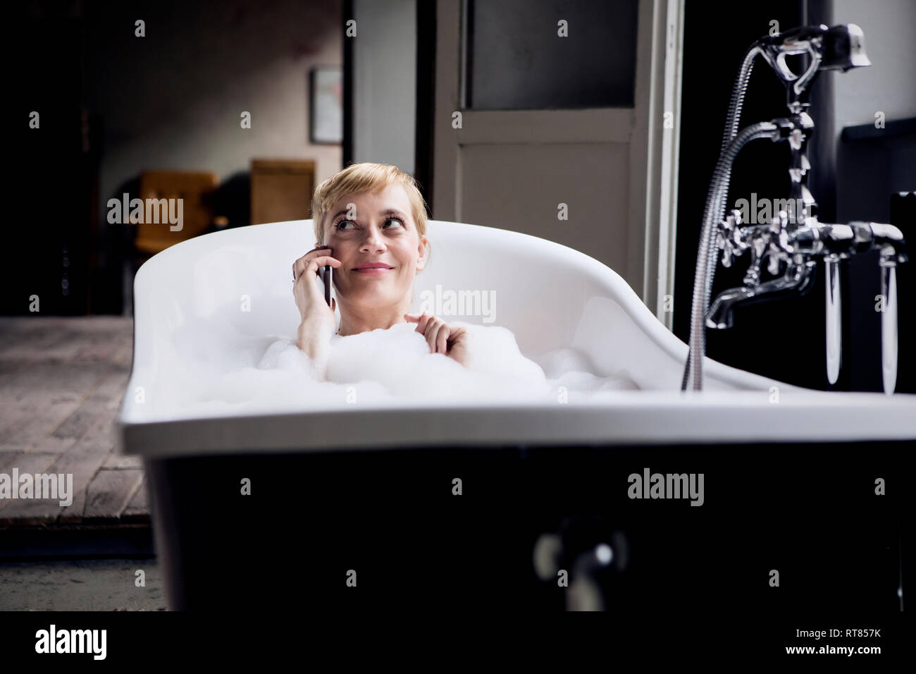 Portrait of smiling blonde femme au téléphone en prenant un bain de mousse dans un loft Banque D'Images