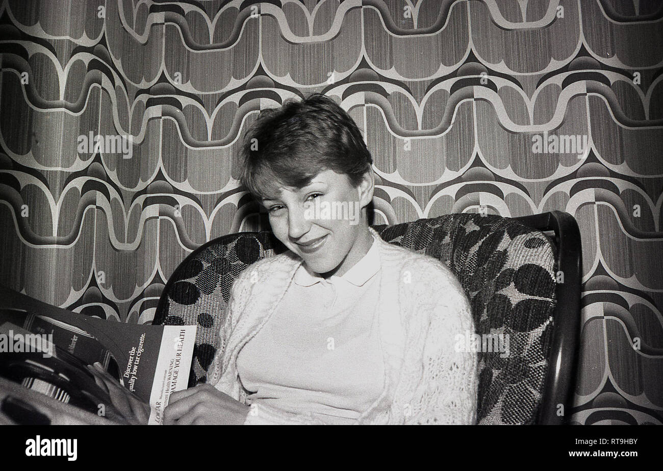 Fin des années 60, historique, attrayant, smiling girl assis sur une chaise en lisant un magazine dans une salle avec le papier peint à motifs de l'époque, Angleterre, Royaume-Uni. Banque D'Images