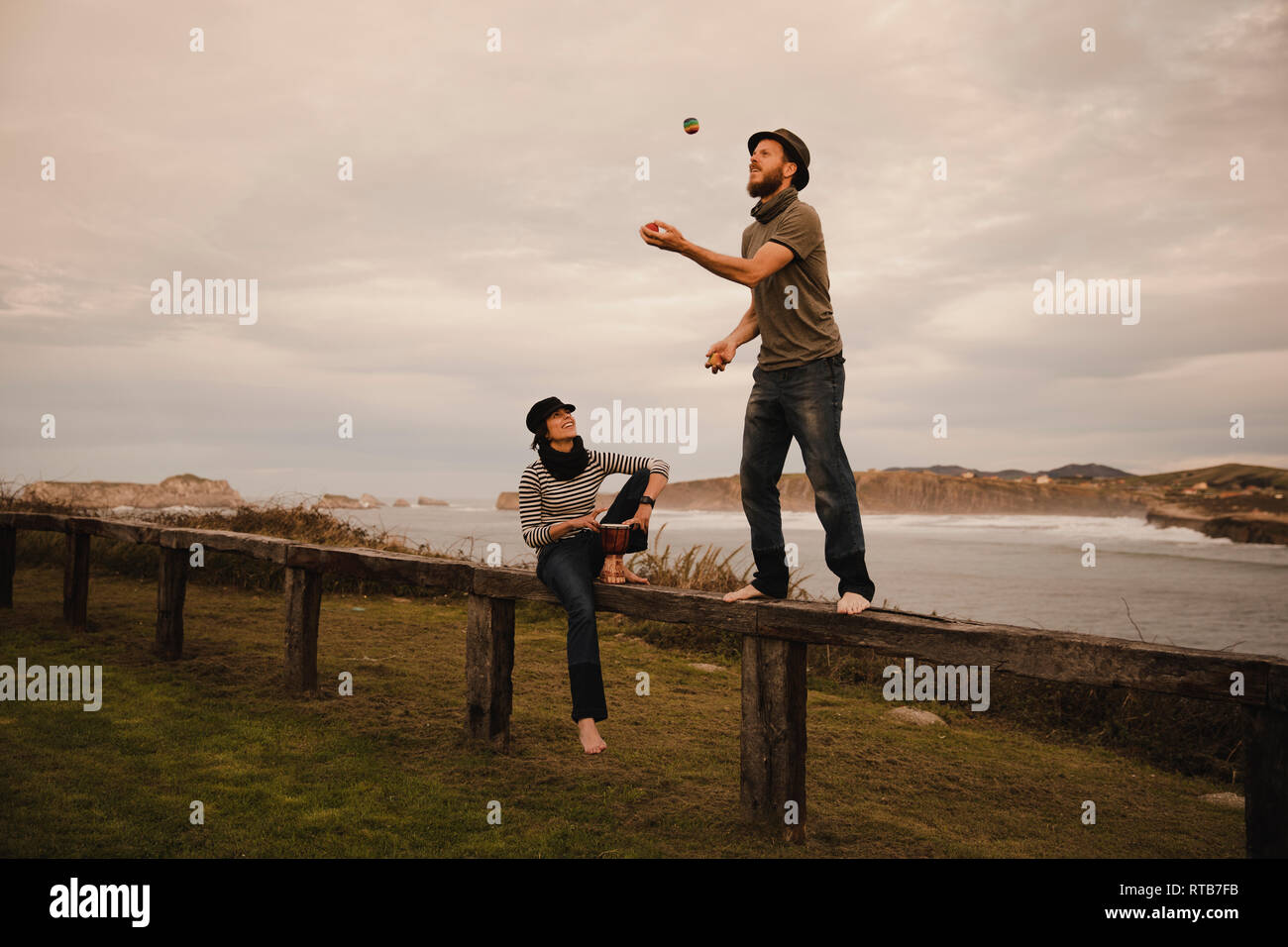 Jeune mec en hat juggling balls près de dame élégante en cap avec tambour d'éthique s'asseoir sur le siège près de la côte de mer et ciel nuageux Banque D'Images