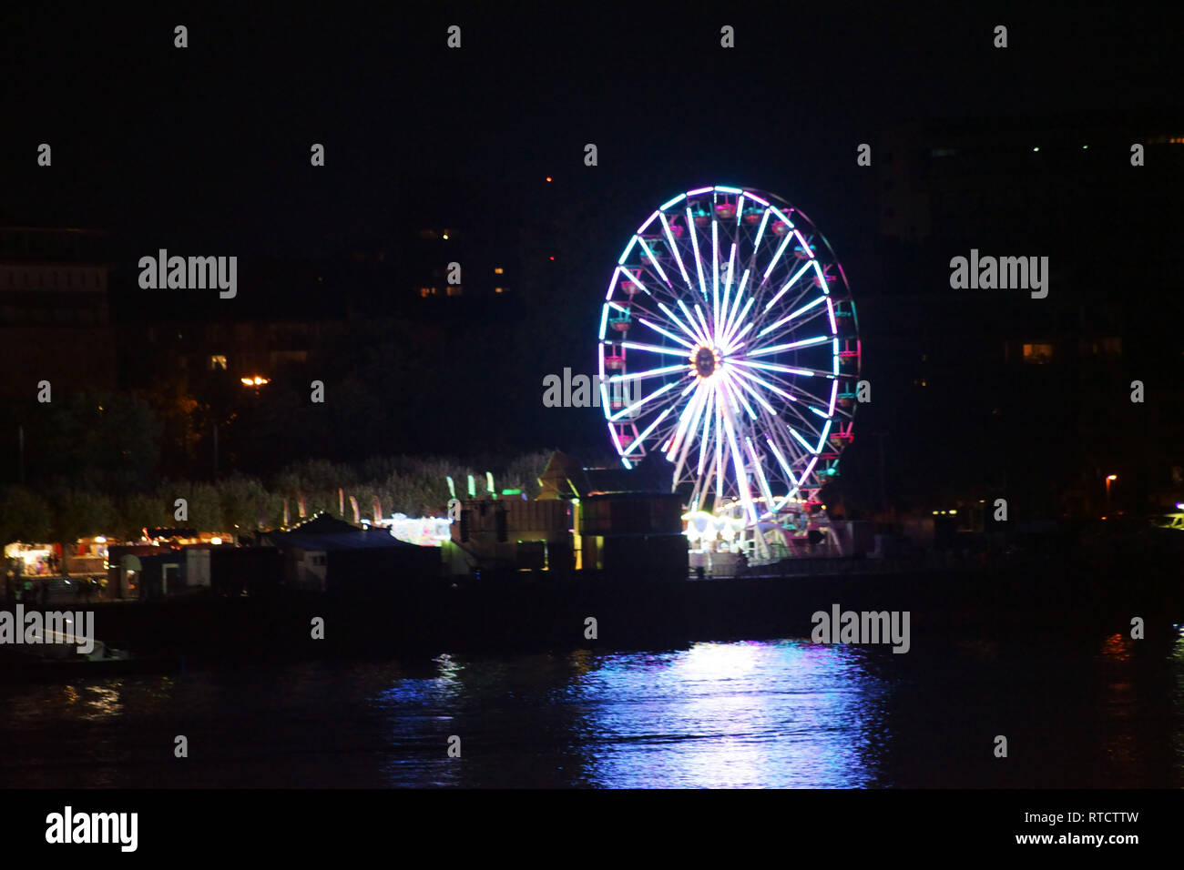 Les lumières d'une grande roue sur une fête foraine dans la nuit derrière une rivière. Banque D'Images