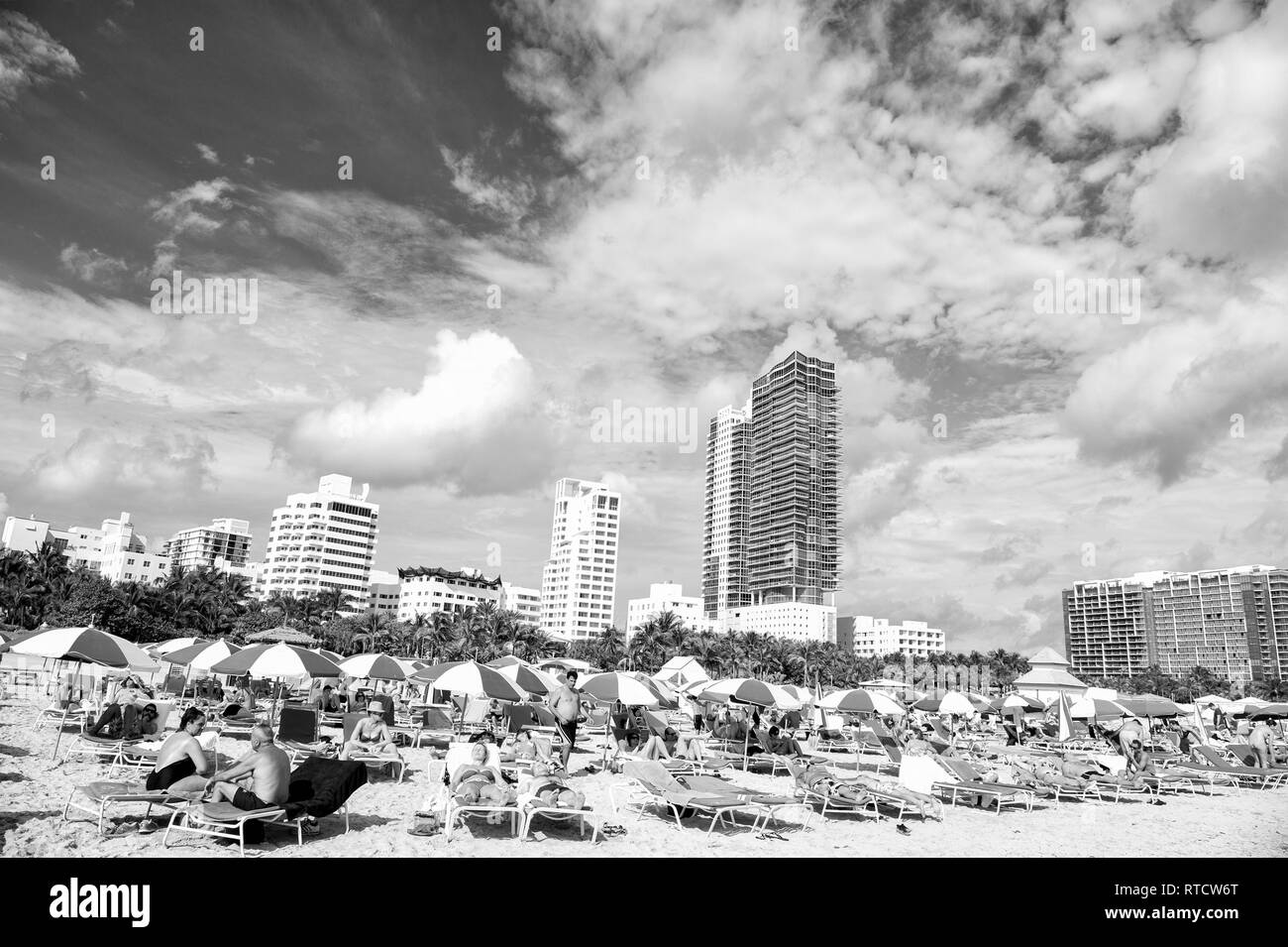 Miami, USA - 10 janvier 2016 : Miami Beach. les gens à prendre le soleil sur des chaises longues sous les parasols de plage bleu avec de hauts bâtiments sur nuageux ciel bleu. Vacances d'été. Lounge et de loisirs Banque D'Images