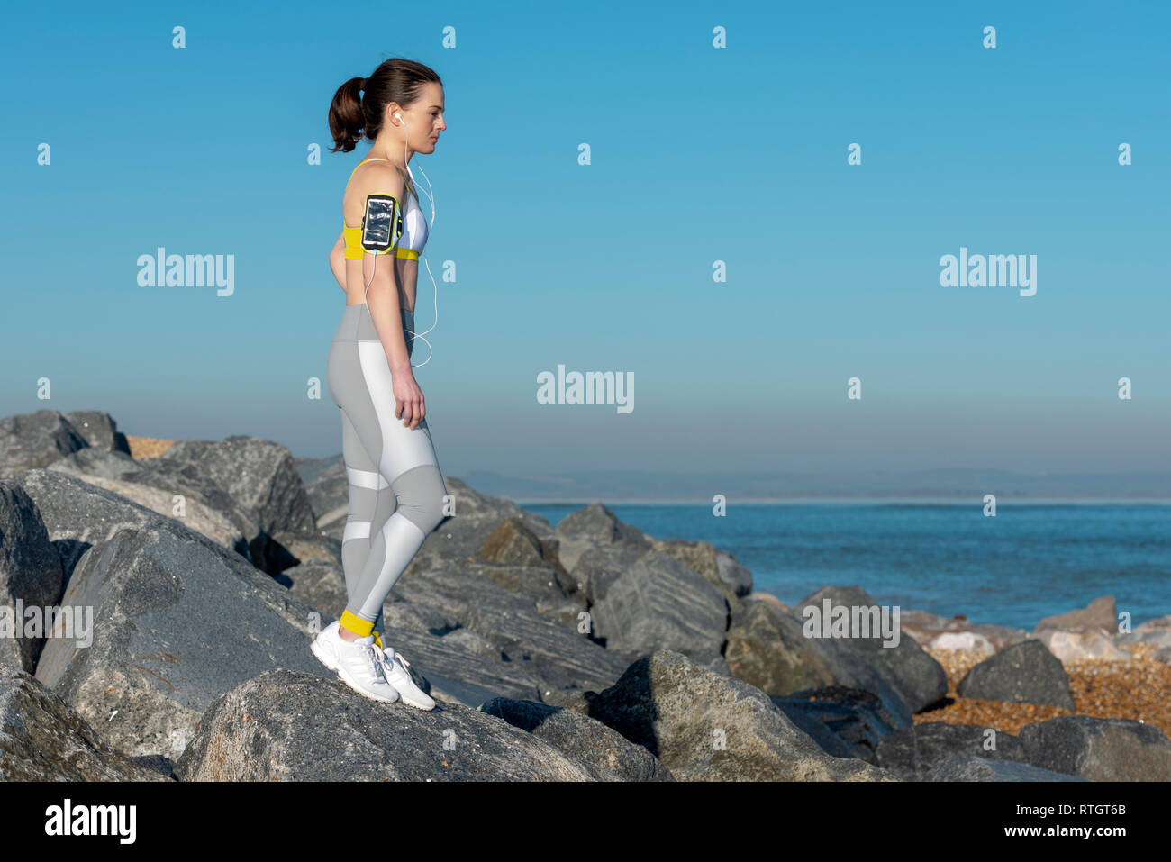Woman wearing sportswear debout sur les rochers au bord de la mer écouter de la musique sur son téléphone sur son brassard. Banque D'Images