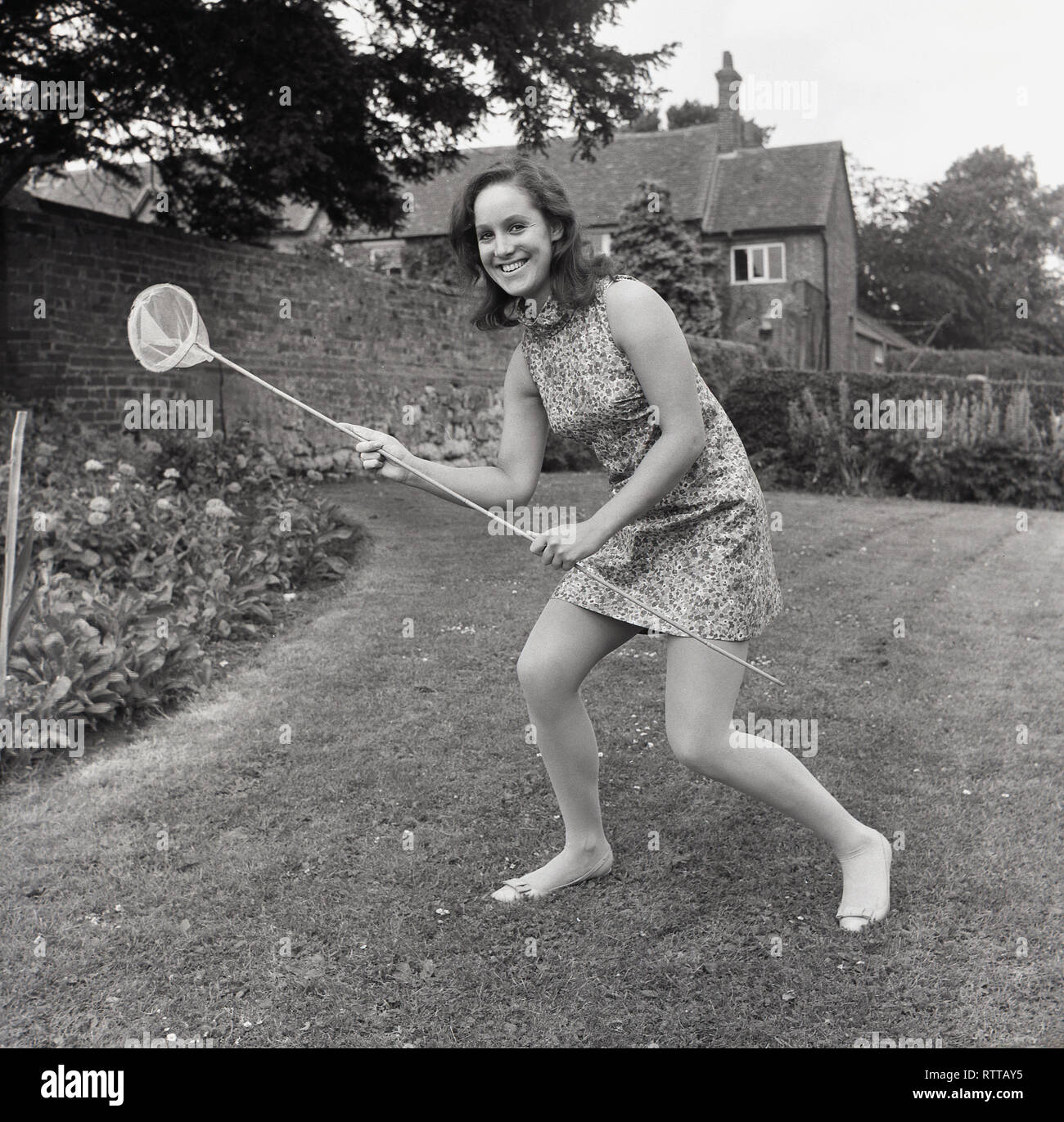 1967, 'flower power'....attraper butterfiles, jeune femme en robe à motifs de fleurs avec un filet de crevettes dans un jardin, England, UK. Banque D'Images