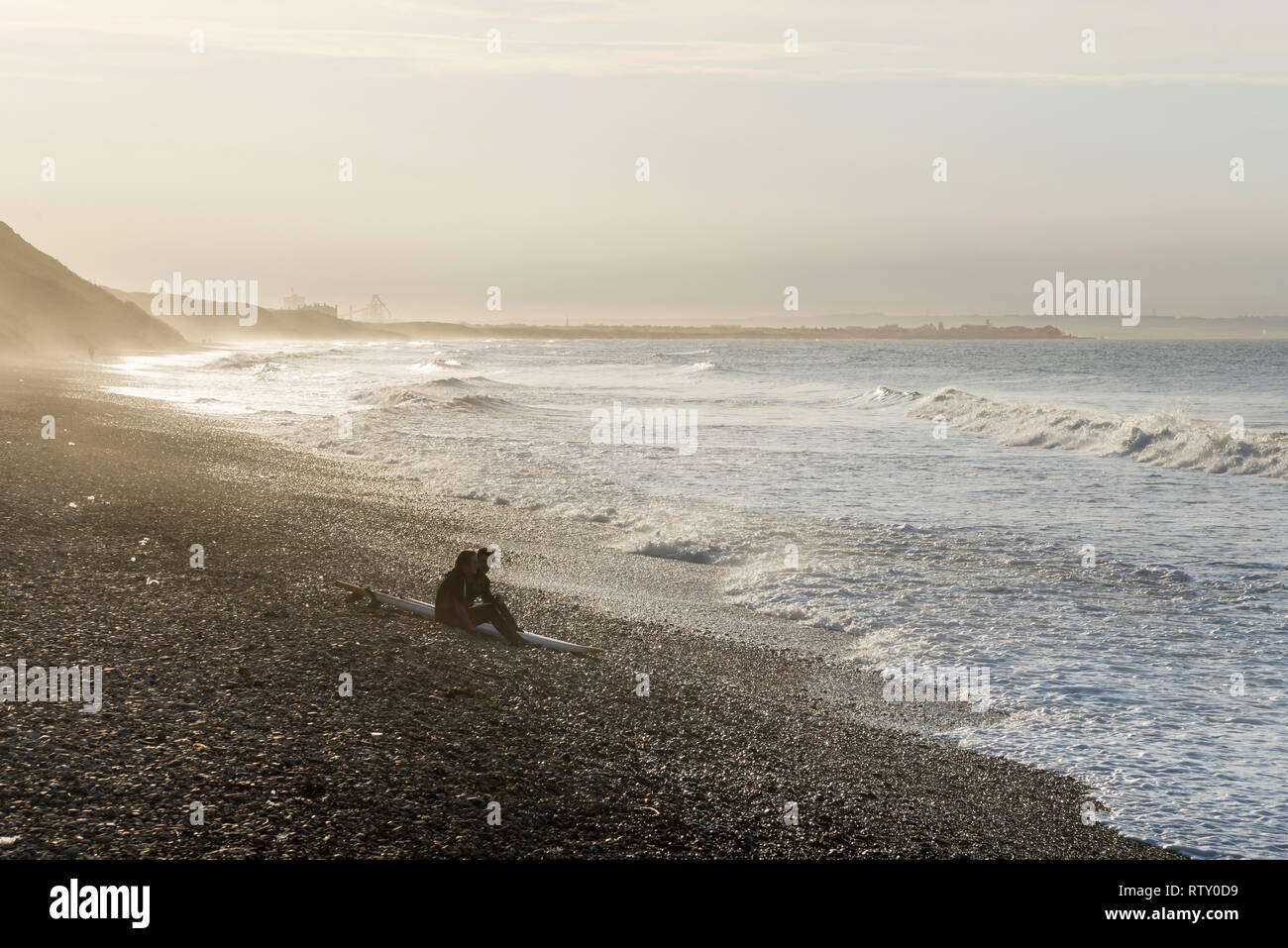 Deux jeunes surfeurs assis sur la plage à Sawai madhopur, North Yorkshire, Angleterre. La lumière du soleil d'or sur un printemps tardif après-midi. Banque D'Images