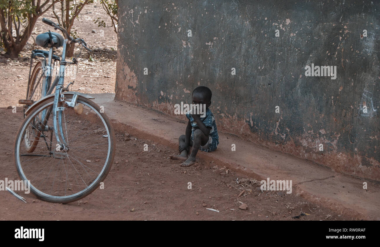 Un jeune garçon assis ghanéen à la grave devant son domicile. Il y a une bicyclette bleue garée à l'extérieur. Banque D'Images