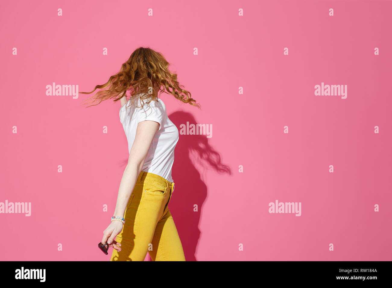 Parti à la mode immersive. Cheerful blonde woman in white T-shirt et jeans jaune branché dansant sur fond rose. Banque D'Images