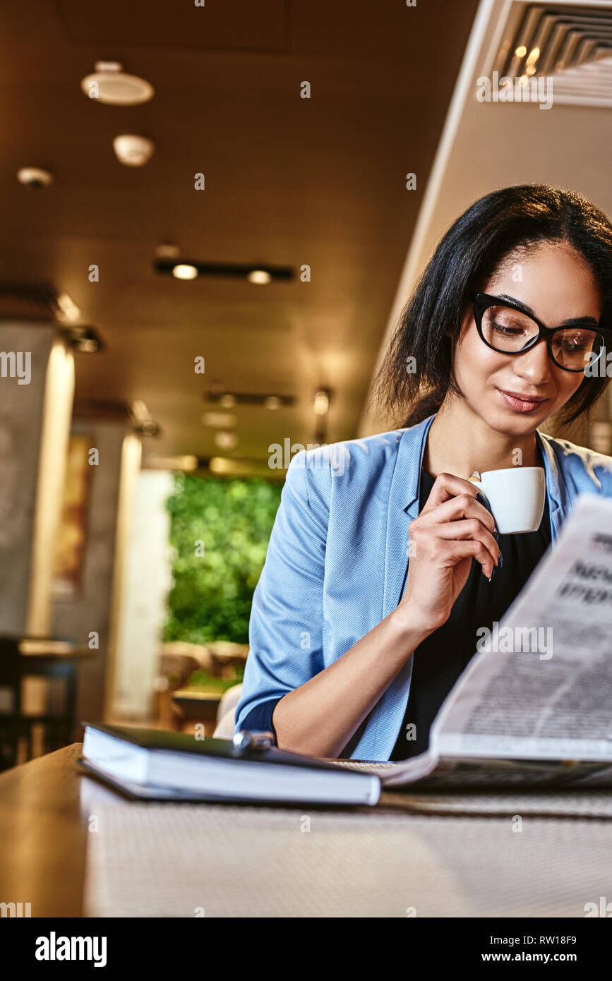 Portrait du jeune chef élégant veste bleu en souriant, tout en regardant à travers des articles dans un journal. Elle est assise à table cafe, boire de morni Banque D'Images